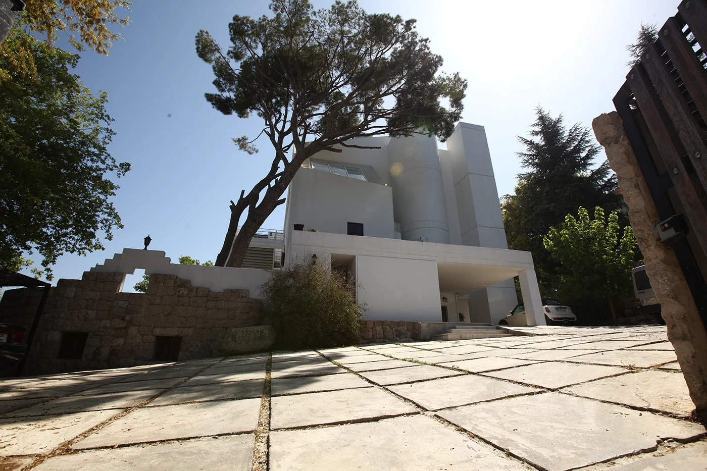 Modern white house with curved and rectangular structures, large tree, stone wall, and parking area in front, under clear blue sky.