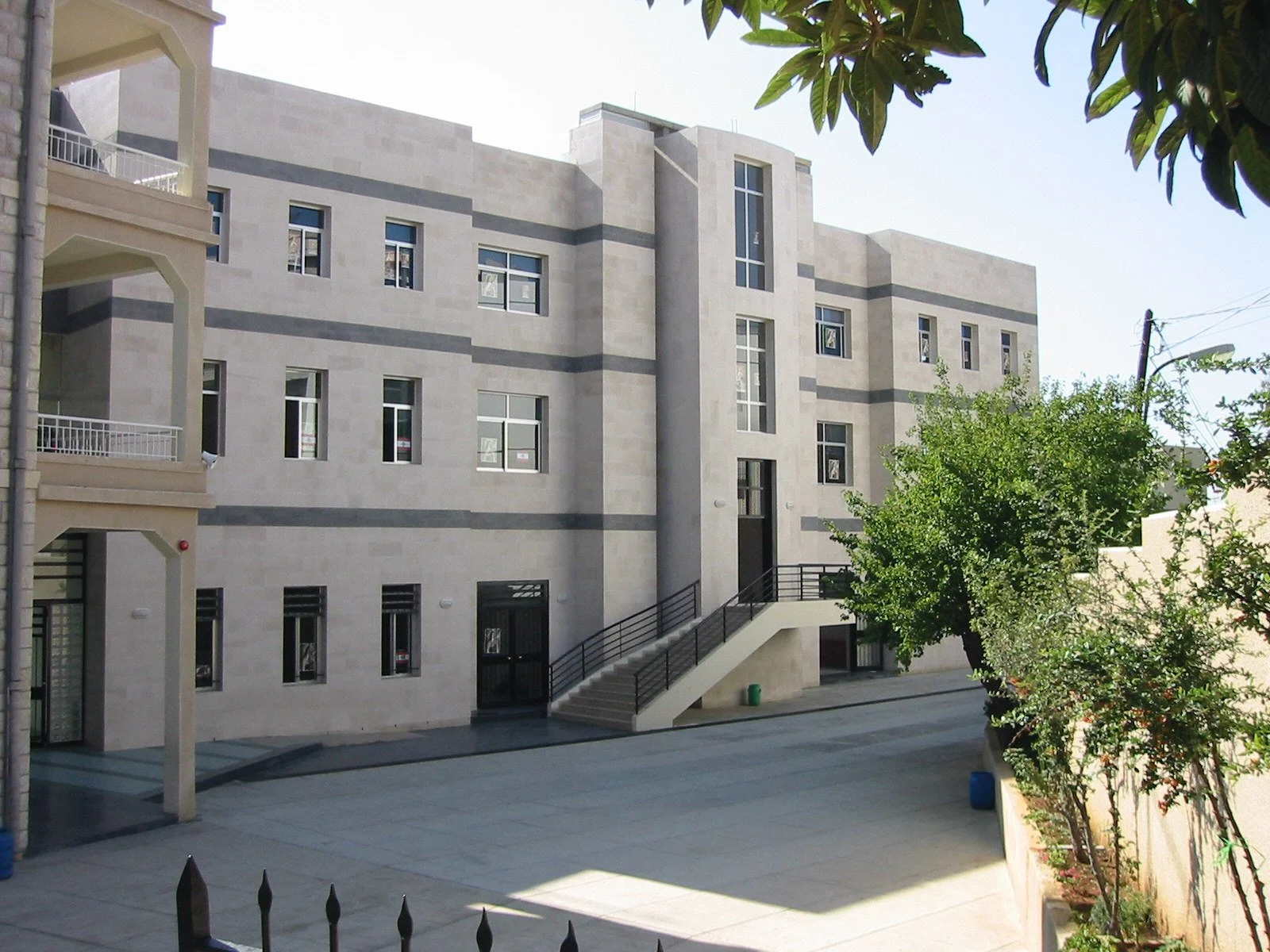 A modern, multi-story building with gray and beige exterior, multiple windows, an external staircase, and a landscaped front yard with trees and shrubs.