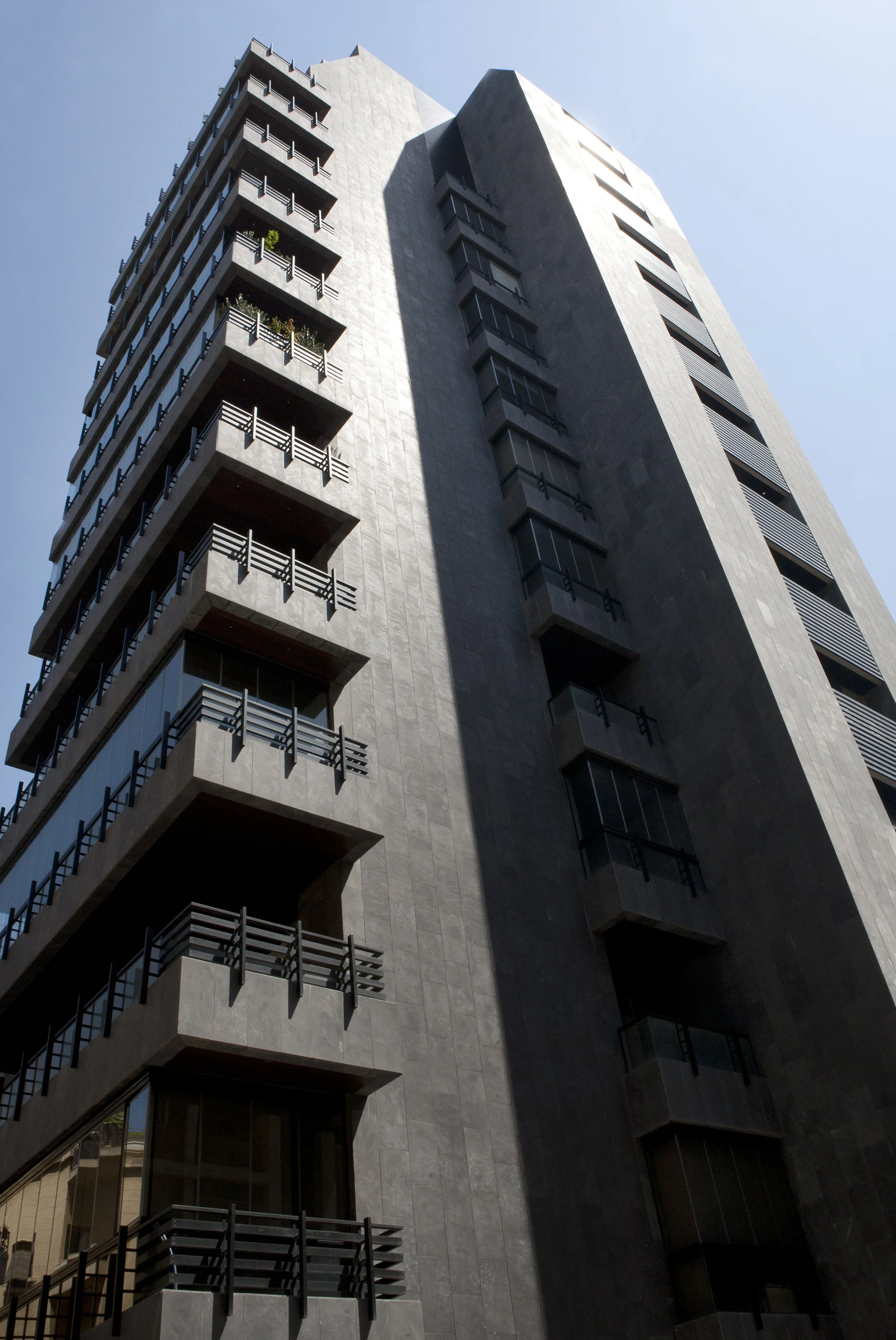 Low-angle view of a modern, dark grey high-rise building with multiple balconies and large windows, under a clear blue sky.