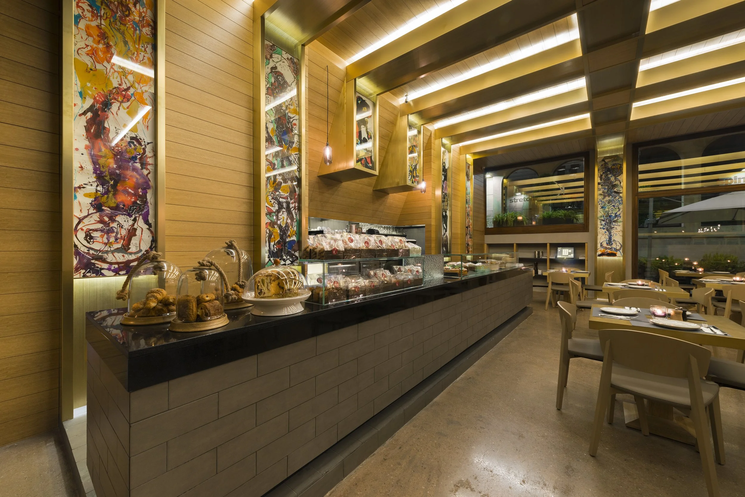 Interior of a modern restaurant with wooden walls and decorative abstract paintings, showcasing a dessert and pastry display counter, tables set for dining, and large windows.