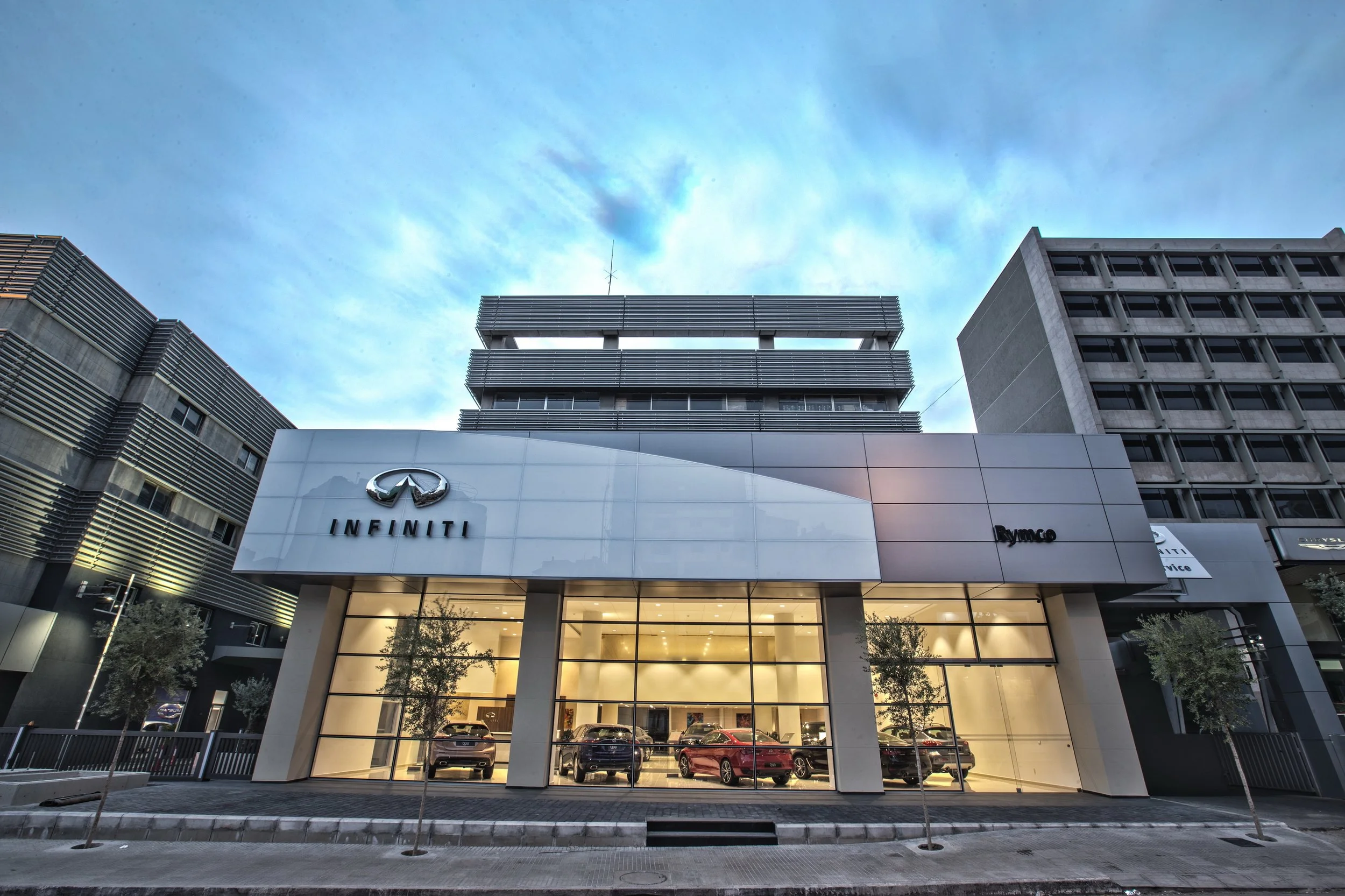 Elevated view of a modern Infiniti car dealership building with a large glass front, showing parked cars inside, and surrounding trees in front. The sky is partly cloudy.