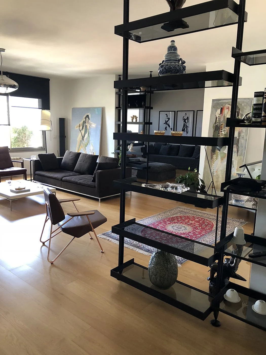 Living room with black sofa, wooden chairs, a white coffee table, and a decorative black shelving unit with vases and small artworks. Artworks and plants are visible on the walls and floor.