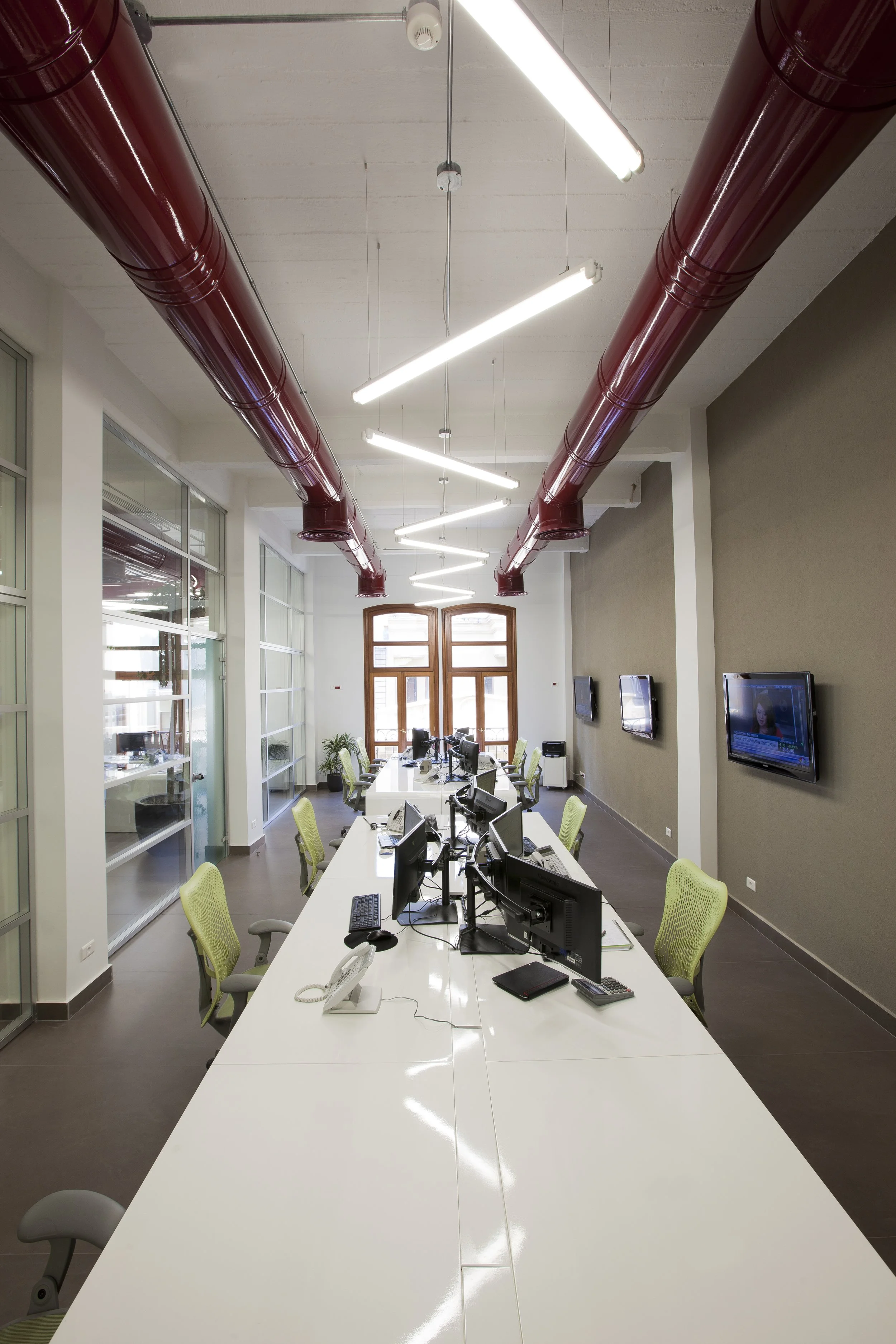 Modern conference room with white tables, green chairs, multiple computer monitors, and wall-mounted TVs, featuring large windows and exposed red industrial pipes on the ceiling.