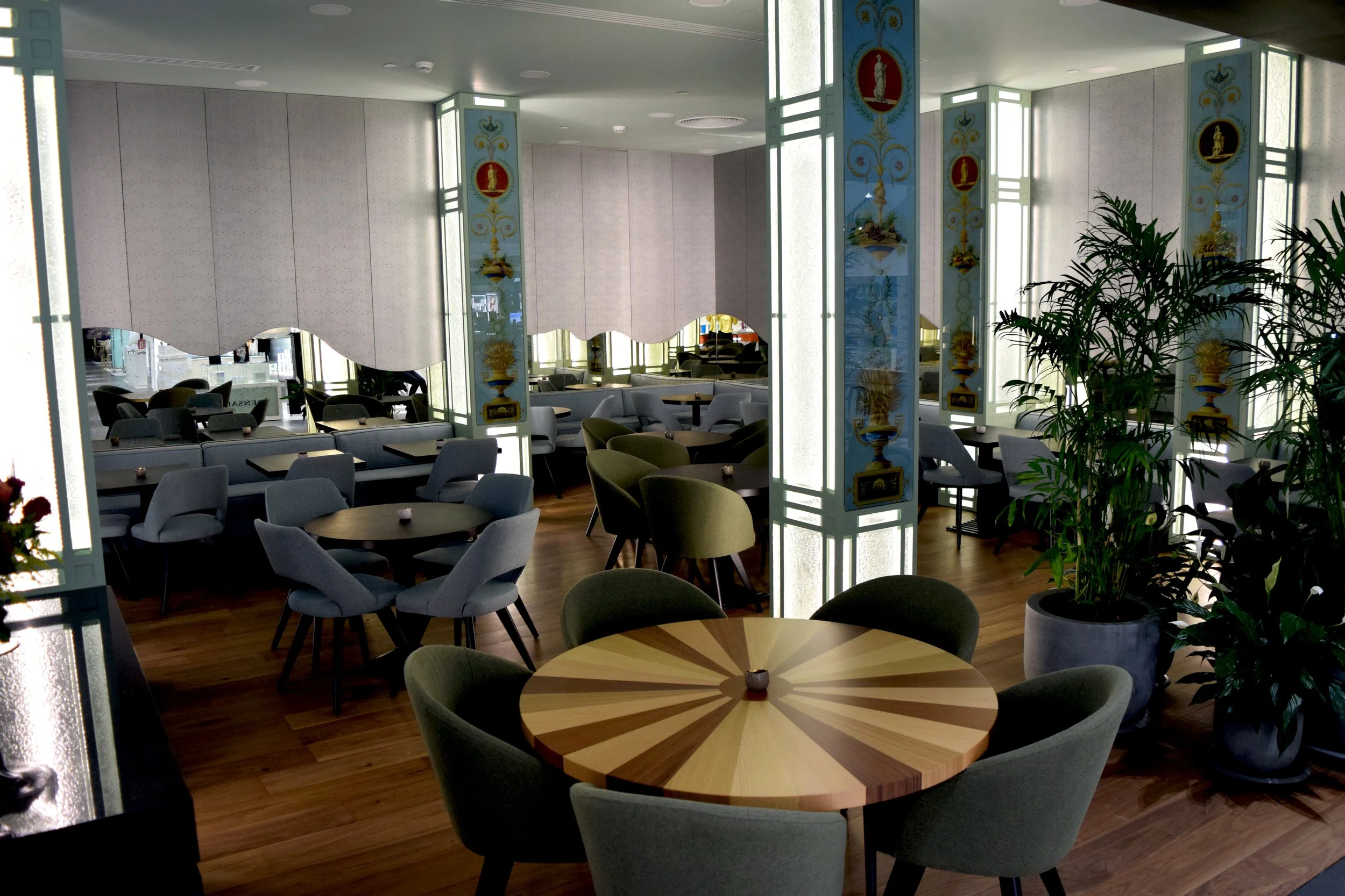 Interior of a restaurant or cafe with round tables, chairs, large potted plants, decorative columns, and sunlit windows.