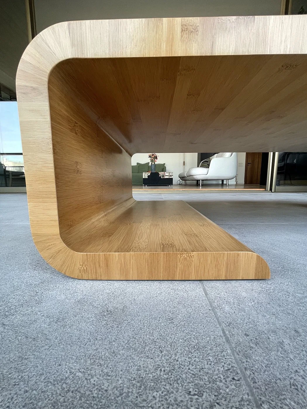 Close-up of a modern wooden coffee table with a hollow center, situated on a gray tiled floor in a living room, with sofa and flowers visible in the background.