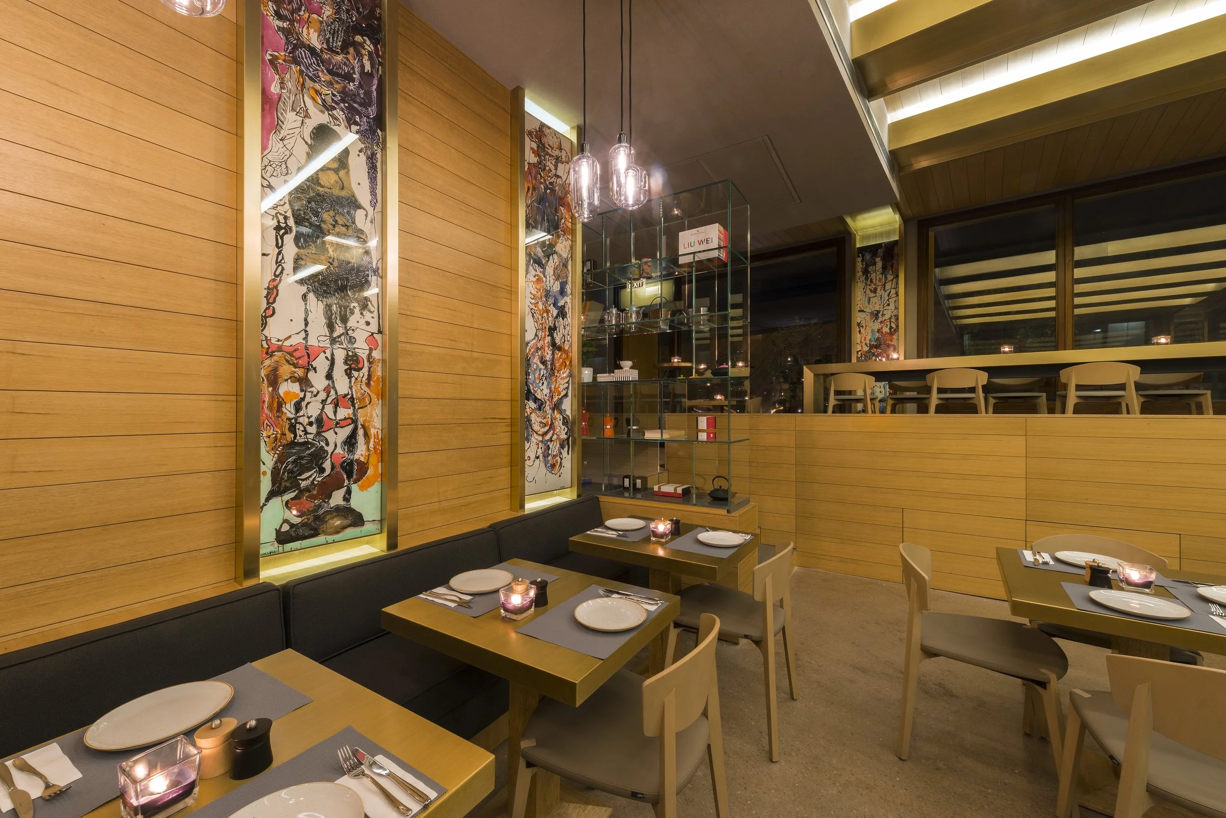 Interior of a modern restaurant with wooden wall panels, abstract art paintings, and candlelit tables set for dining. There are large windows and hanging light fixtures.