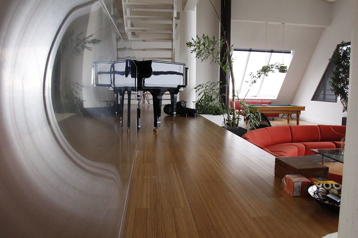 Interior of a modern living space with a polished piano, a red sectional sofa, potted plants, and large skylight windows.