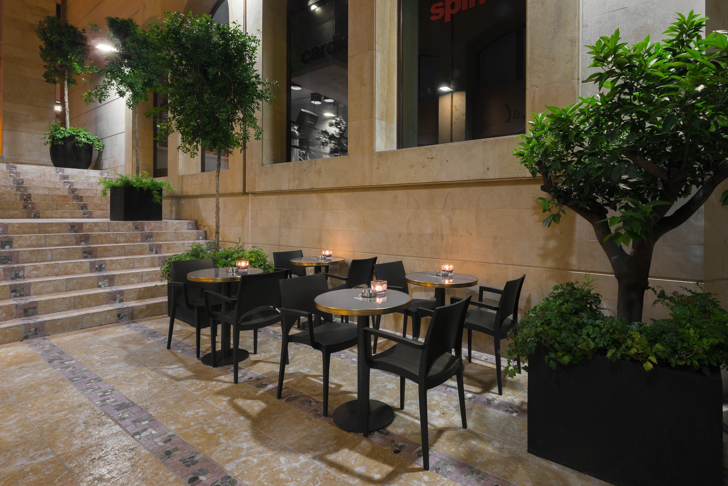 Outdoor dining area with black tables and chairs, surrounded by green potted trees and plants, with a staircase and a beige stone building in the background.