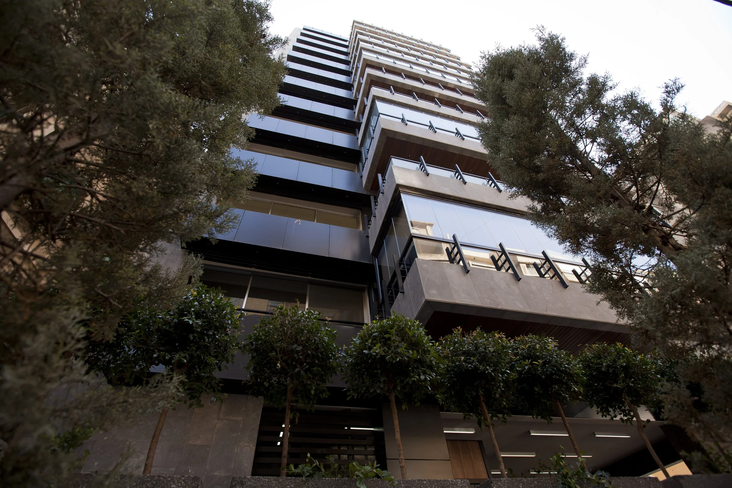 Low-angle view of a tall modern multi-story building with glass windows and balconies, flanked by trees in the foreground.