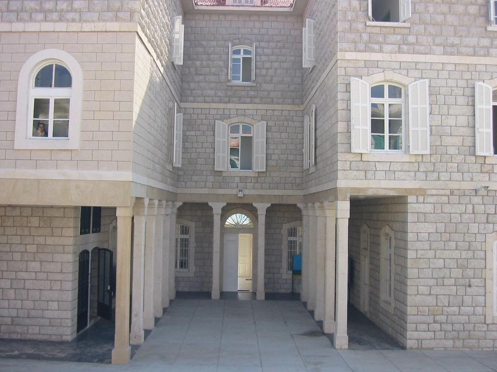 View of a large stone building with multiple windows and white open shutters, features arches and columns at the entrance, with a paved walkway leading to the door.