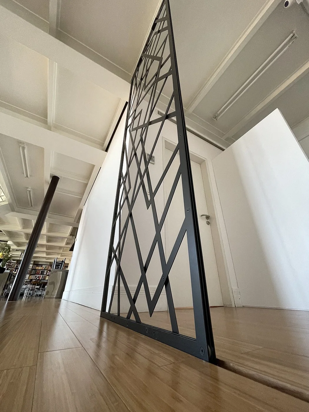 A black metal decorative grille or room divider with geometric patterns, placed on a wooden floor in an interior space.