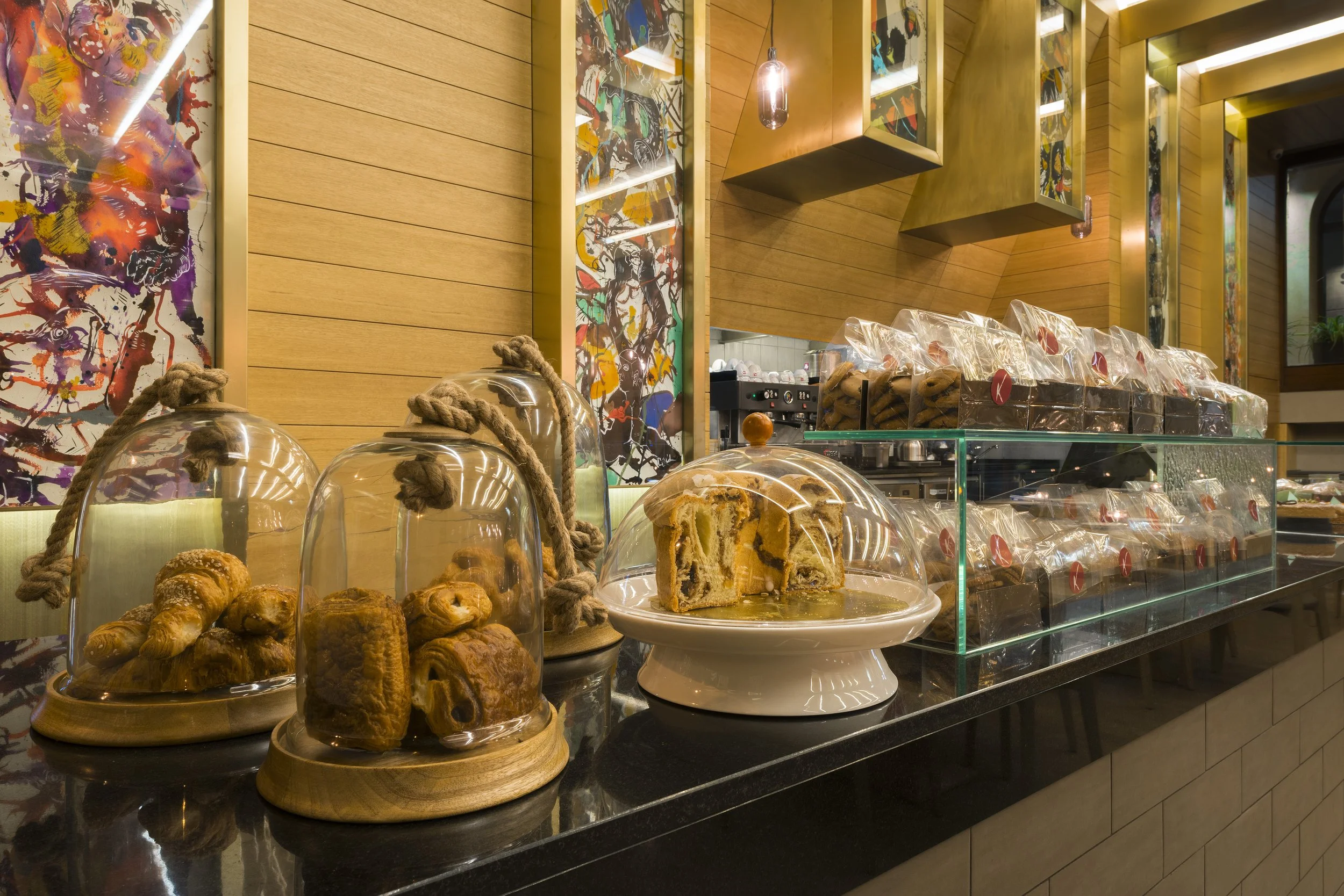 A bakery display with various pastries under glass domes, including croissants and bread, set on a black countertop with a wooden background and colorful abstract artwork on the wall.