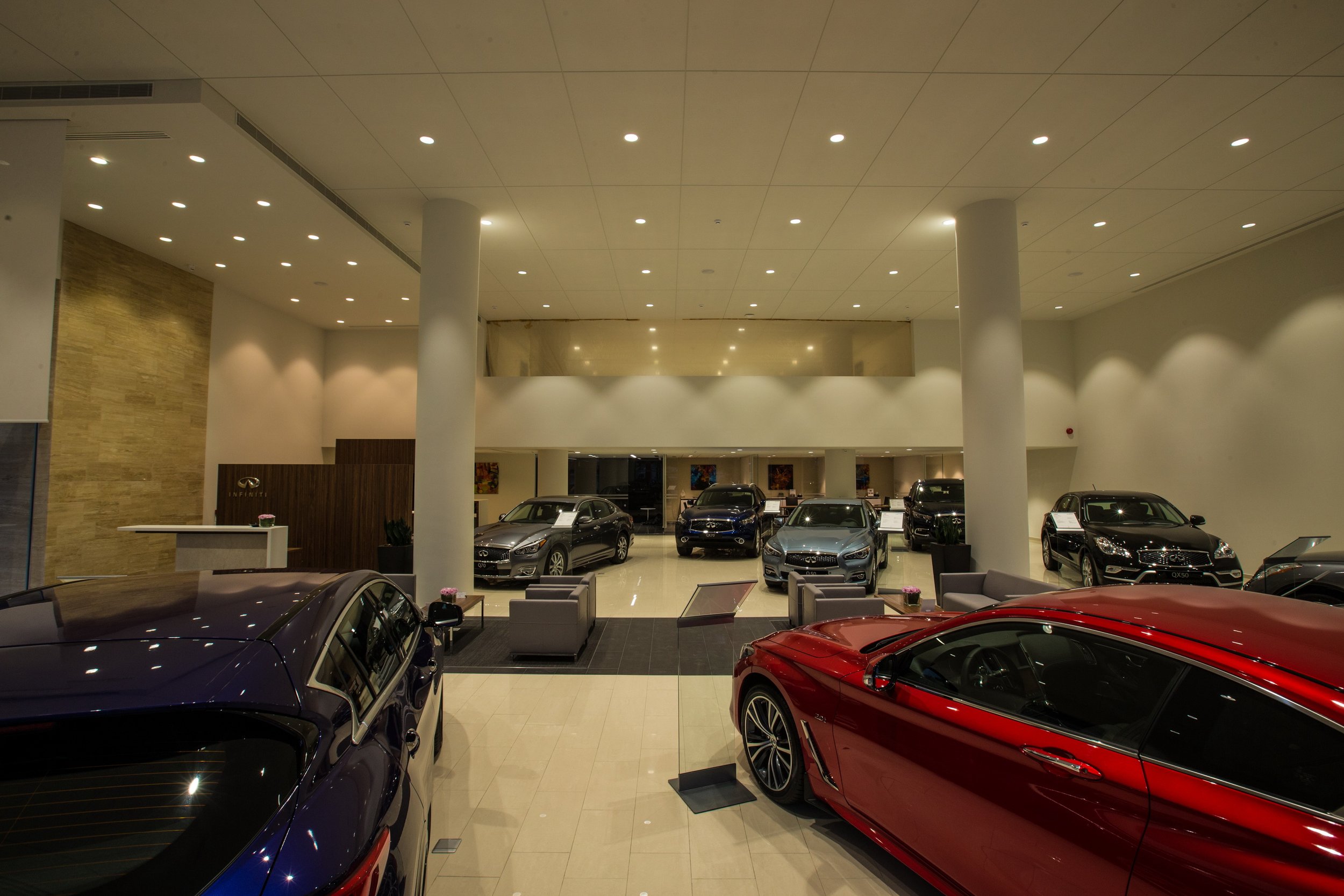 Inside a luxury car dealership showroom with various cars on display, high ceilings with recessed lighting, and a seating area.