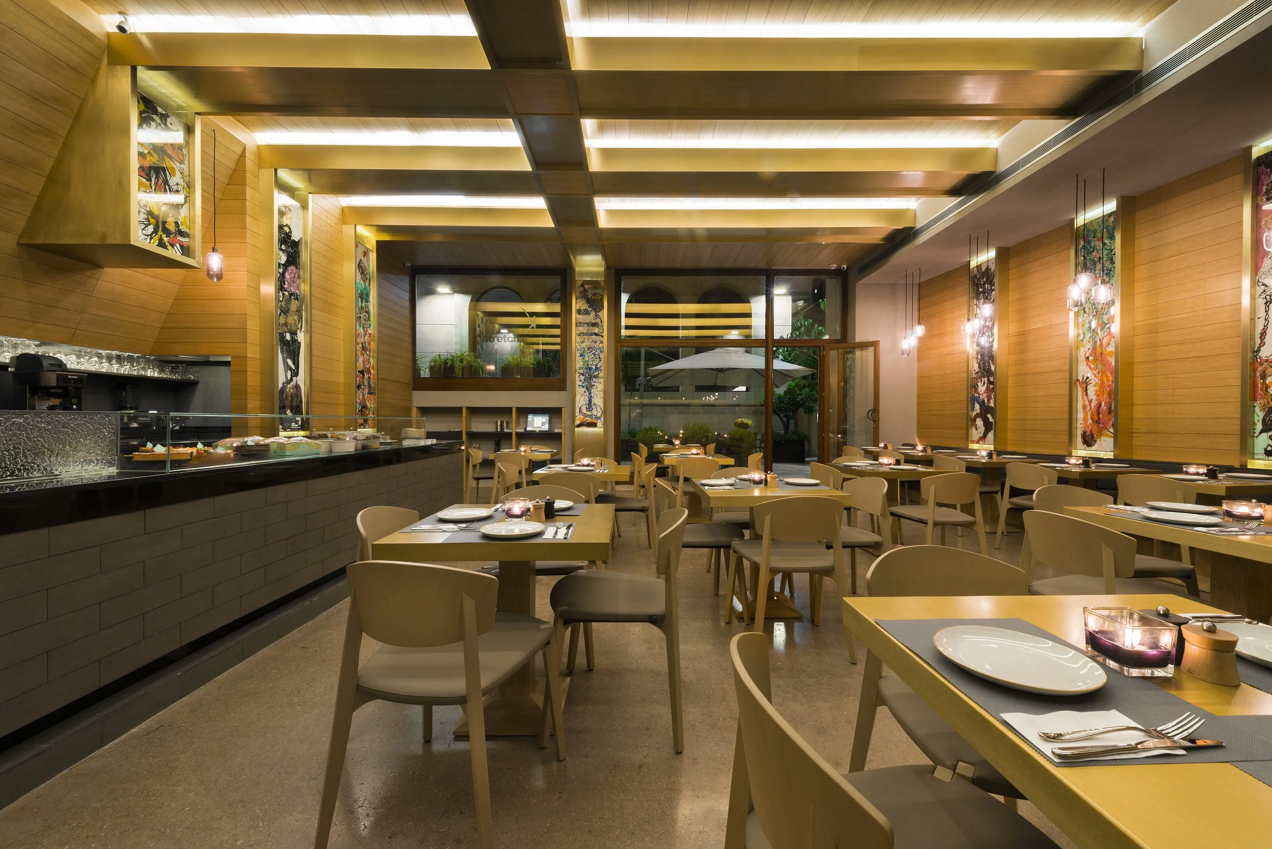 Modern restaurant interior with wooden walls, hanging pendant lights, neatly set tables with plates and cutlery, and large windows showing outdoor greenery.