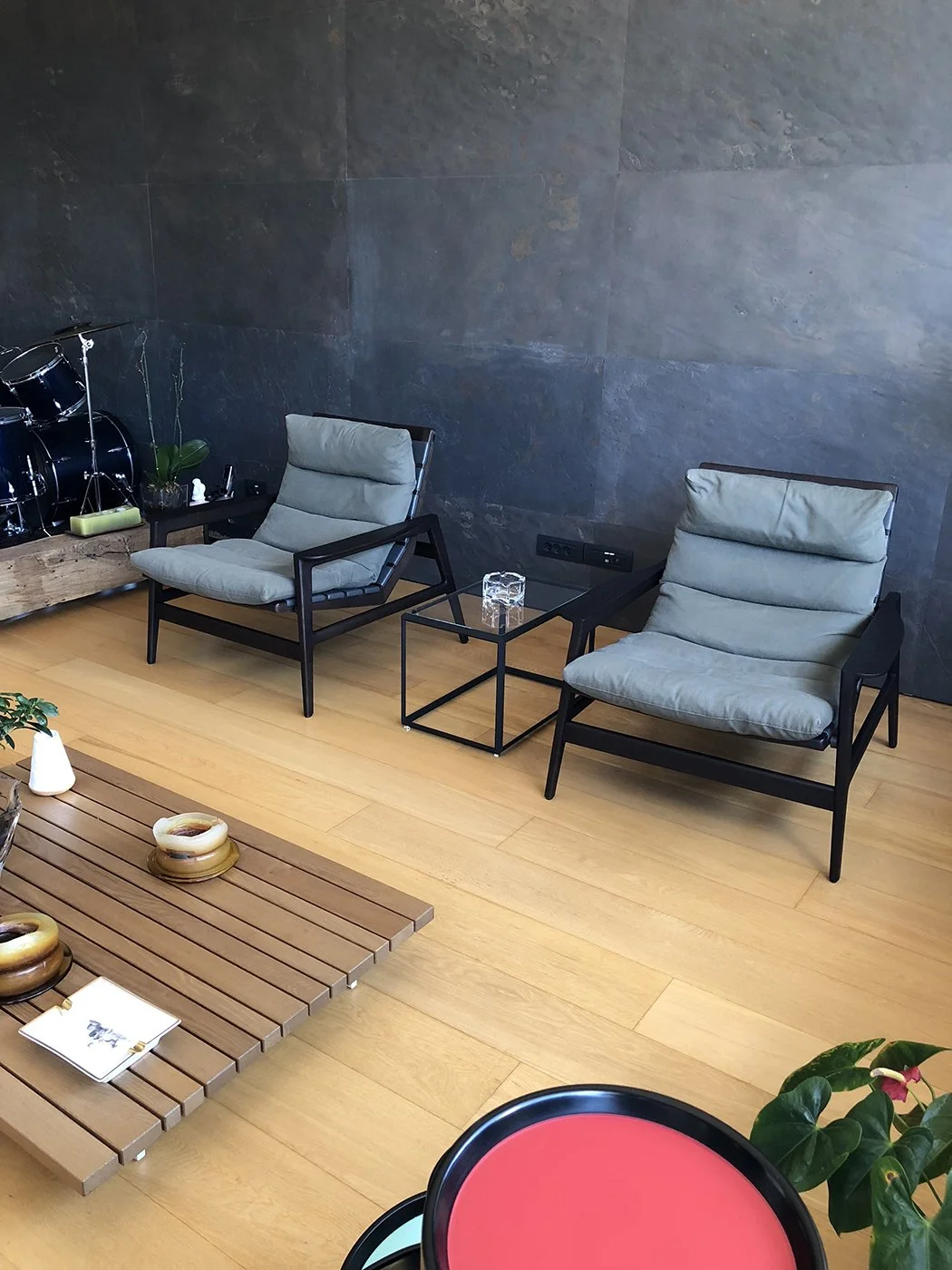 Two modern lounge chairs with light gray cushions, a small black metal side table with a glass top holding a glass container, and a black wall socket on a dark gray textured wall in a cozy indoor space with wooden flooring.