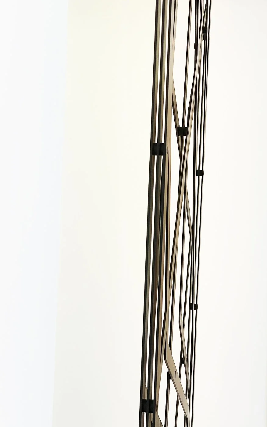 Close-up of a modern, minimalist black metal sculpture or structure with intersecting vertical and diagonal lines against a plain white wall.