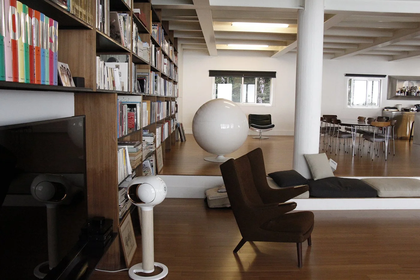 Interior of a modern room with wooden floors, a large bookshelf filled with books, a white spherical lamp, and various chairs and seating areas, including a brown chair, a beige chaise lounge, and a black swivel chair. Multiple windows allow natural 