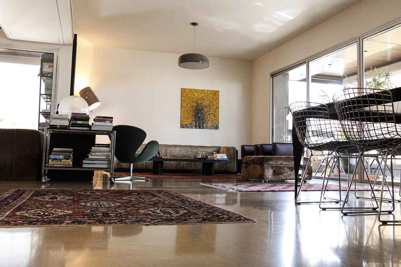 Living room with large windows, an area rug, a sectional sofa, a dining table with wire chairs, books, and artwork on the wall.