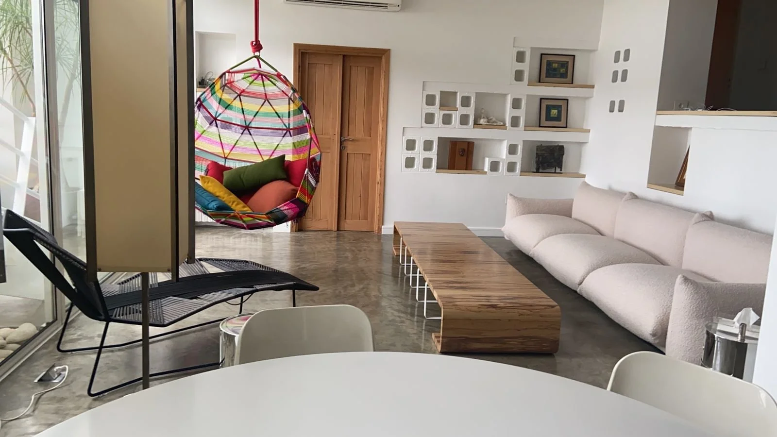 Living room with beige sofa, wooden coffee table, colorful hanging chair, and built-in white shelves on the wall.