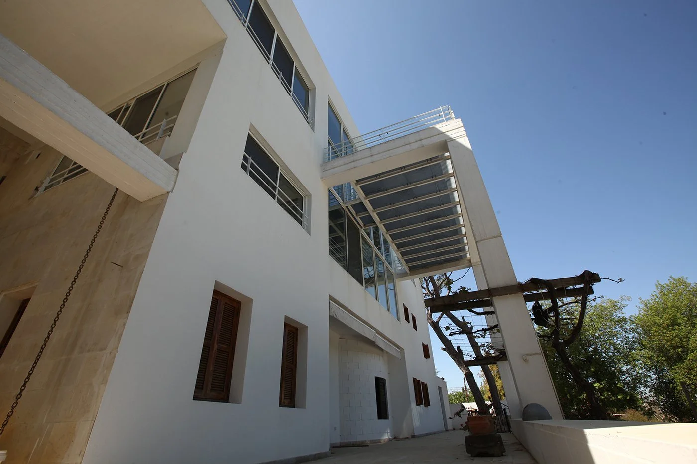 Modern white multi-story building with glass windows, metal balconies, and a metal sunshade, next to a tree with green leaves against a clear blue sky.