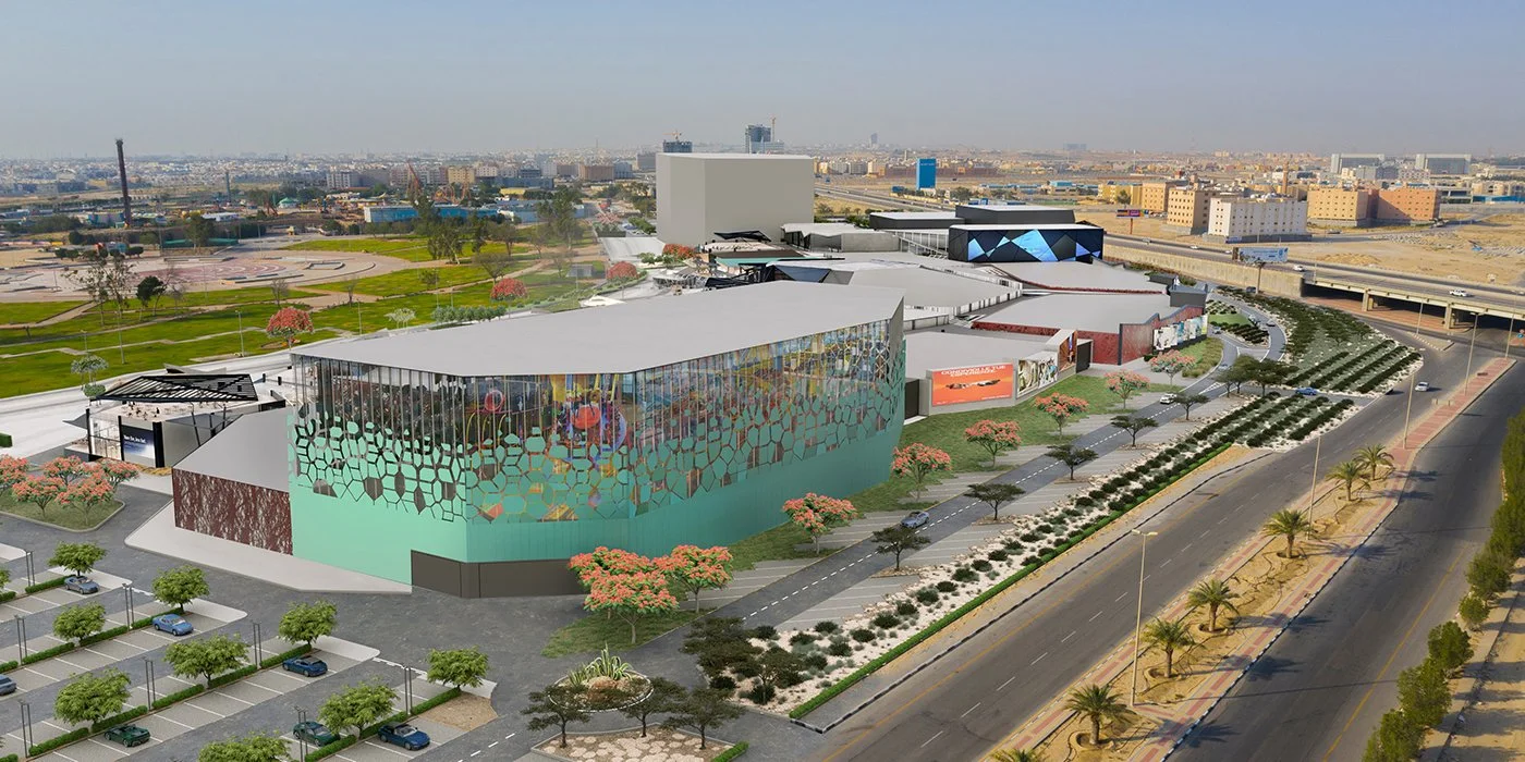 A large, modern shopping mall with colorful exterior and glass walls, surrounded by parking lot with cars and landscaped greenery, in an urban setting.
