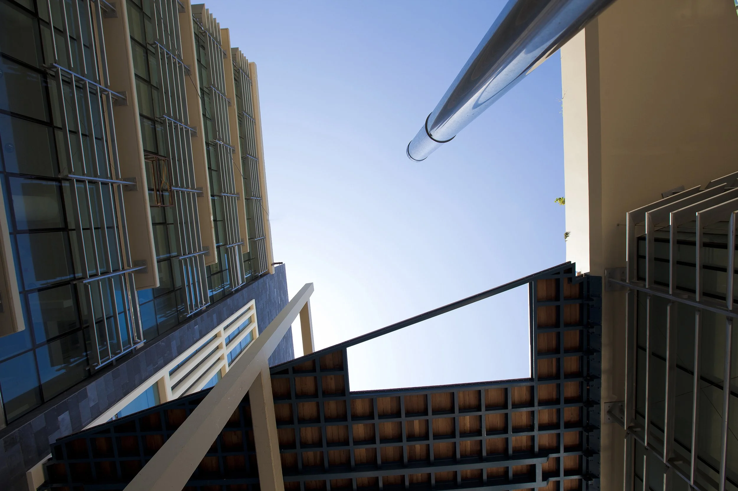 Looking upward at the clear blue sky through the open courtyard of a modern multi-story building, showing glass windows, metal railings, and architectural details.