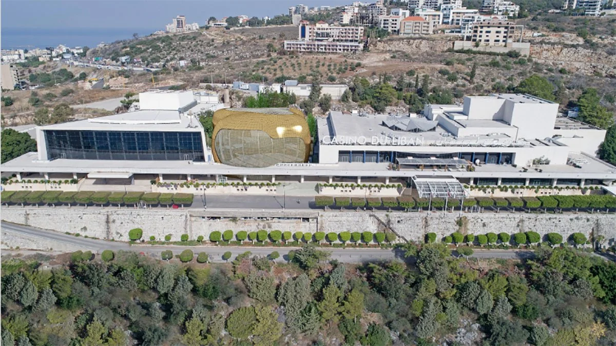 Aerial view of Casino Dubai, a modern building with a gold and glass facade, situated on a hillside with trees and greenery below and urban buildings in the background.