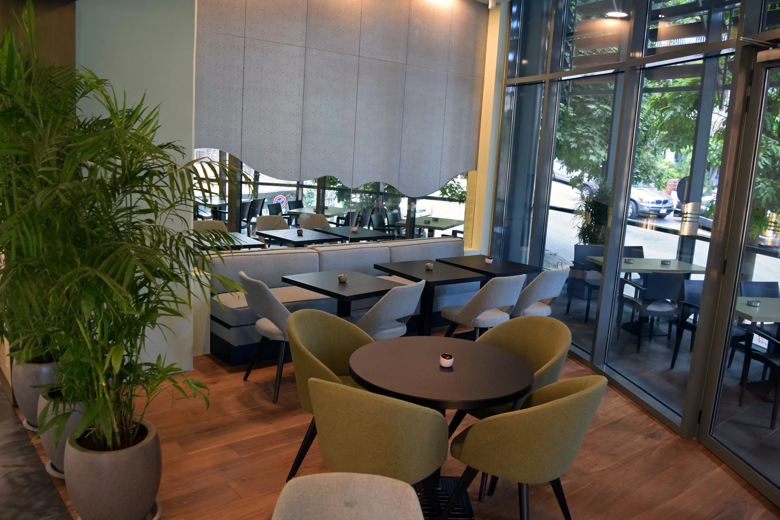 Interior of a modern restaurant or café with tables and chairs next to large glass windows, a potted plant in the foreground, and a mirror on the wall.