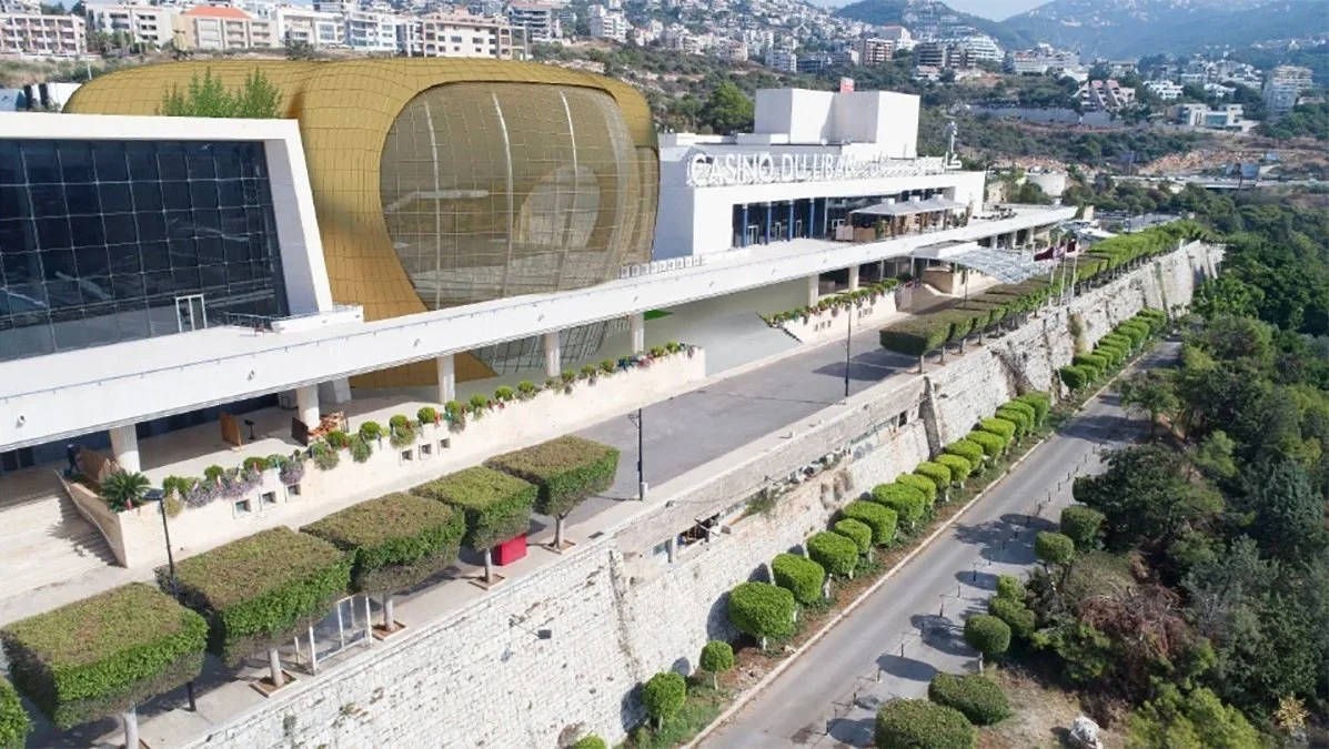 Aerial view of a modern building with a rounded gold facade, glass windows, and outdoor terrace, surrounded by neatly trimmed trees and bushes on a hillside with a cityscape in the background.