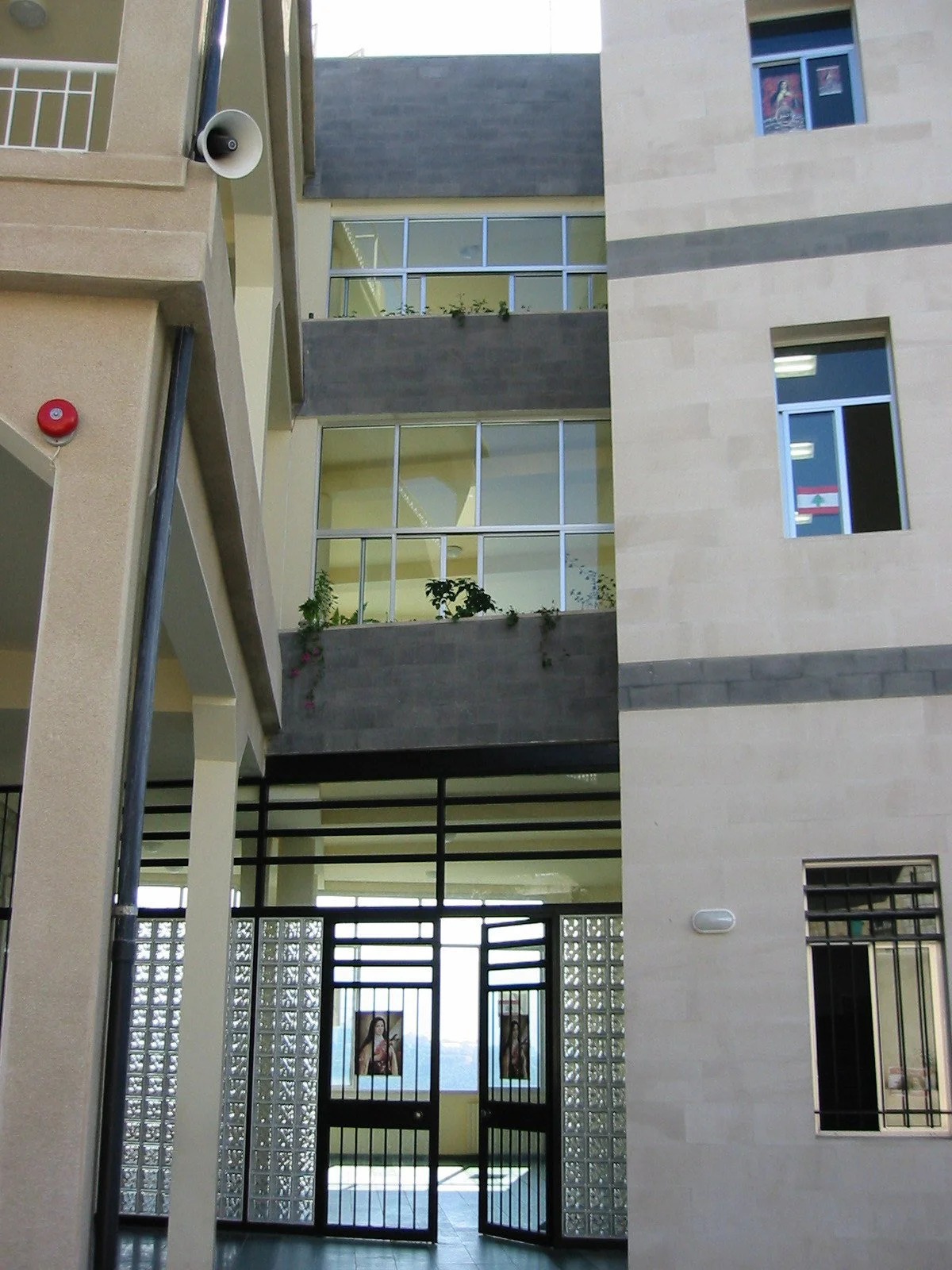 View of a multi-story building's interior from the ground floor, showcasing balconies with glass railings and potted plants, along with windows and doors, including a gate with religious images.