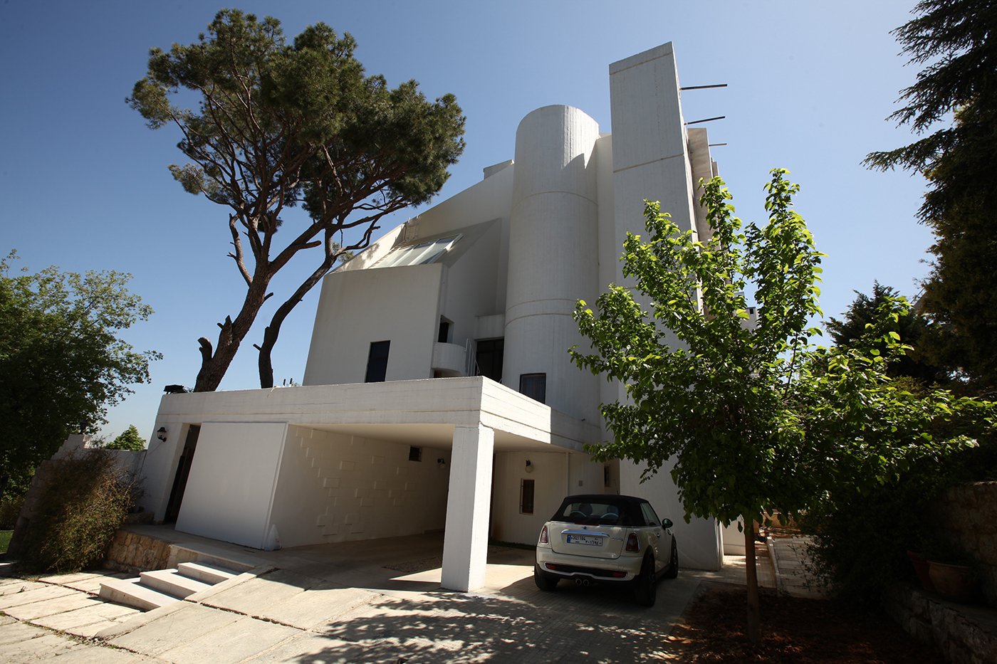 Modern white residential building with curved and rectangular architectural elements, a car parked underneath a shaded carport, and trees in the foreground and background, under a clear blue sky.