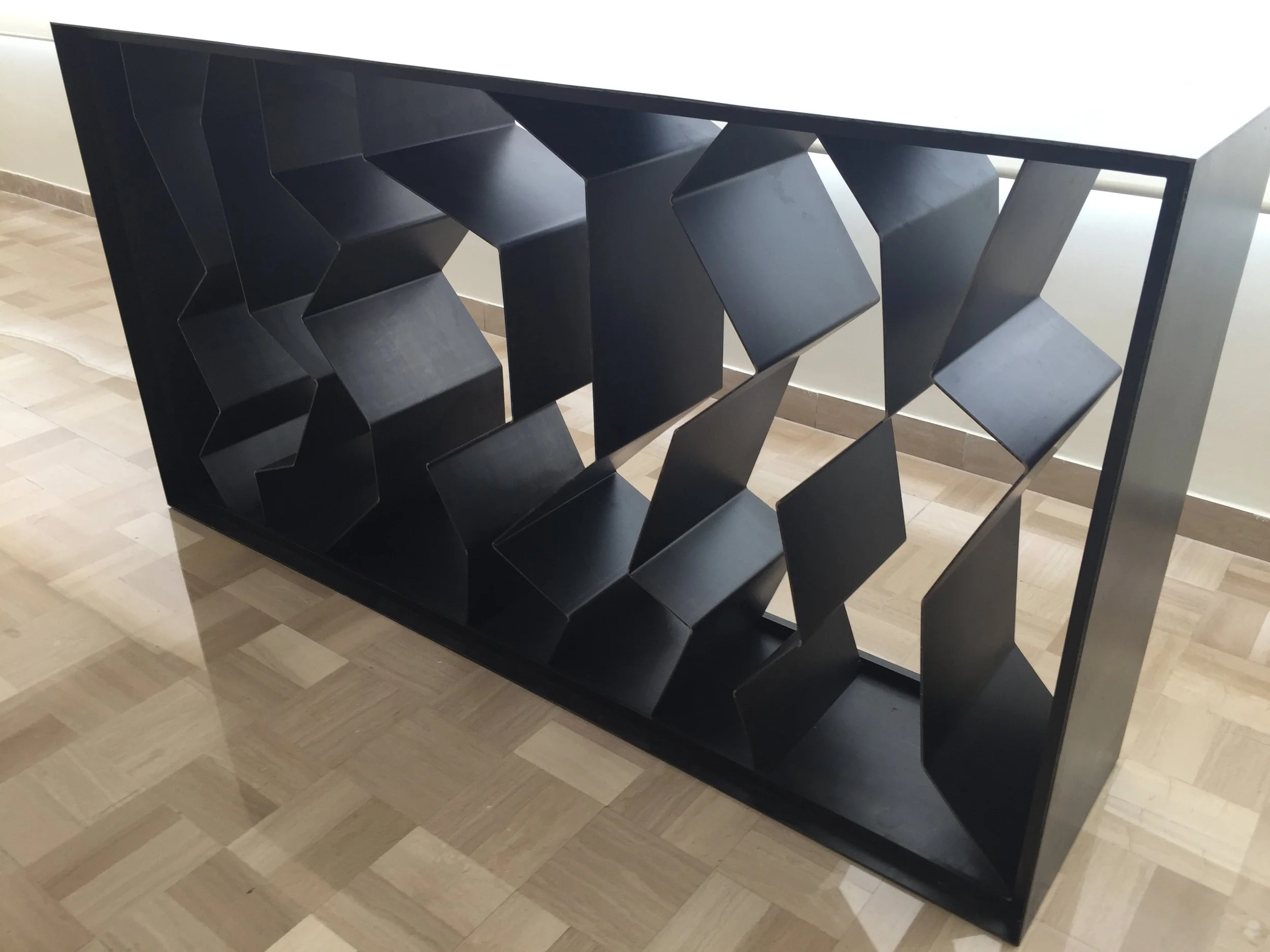 A modern black bookshelf with geometric, zigzag-shaped shelves, placed against a white wall and on a wooden floor.