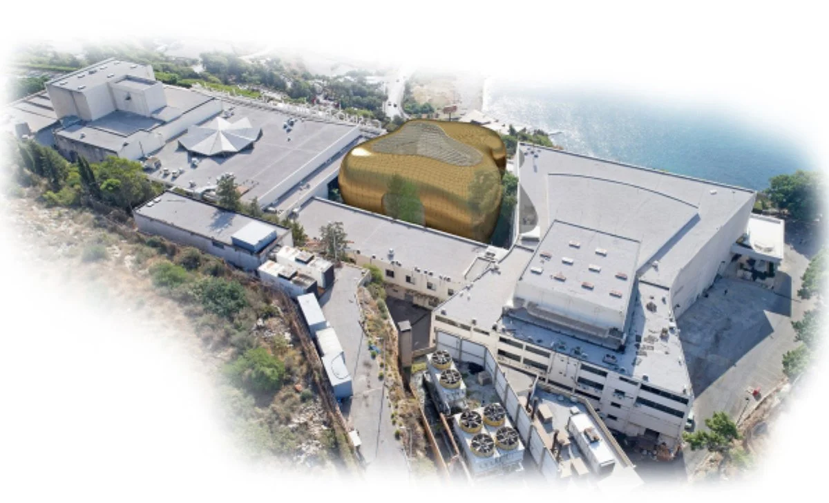 Aerial view of a large building complex with a gold-colored, irregularly shaped structure and adjacent white buildings near water, with some green vegetation around.