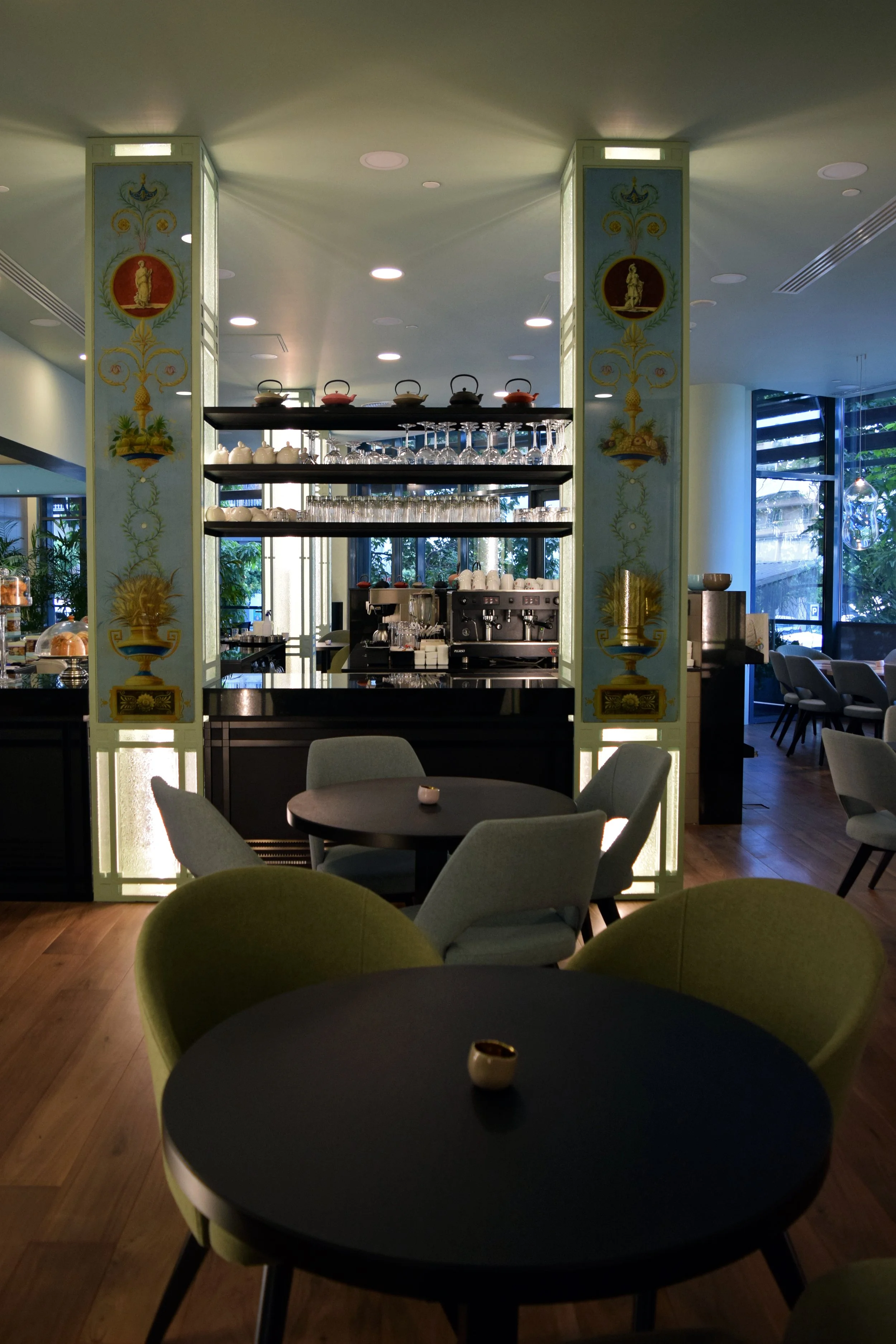 Inside a modern restaurant or cafe with green and black chairs, round tables with small white cups, and a bar area with shelves of glassware and cups, illuminated by ceiling lights and natural light through large windows.