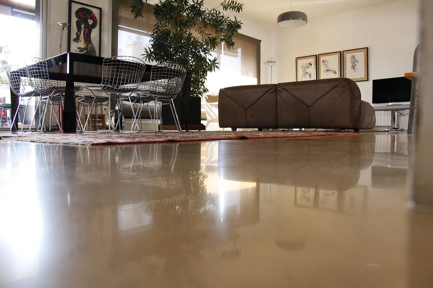 Living room with dining table, chairs, brown leather sofa, artwork, TV, large window, potted plant, and hanging ceiling light, reflected on glossy floor.