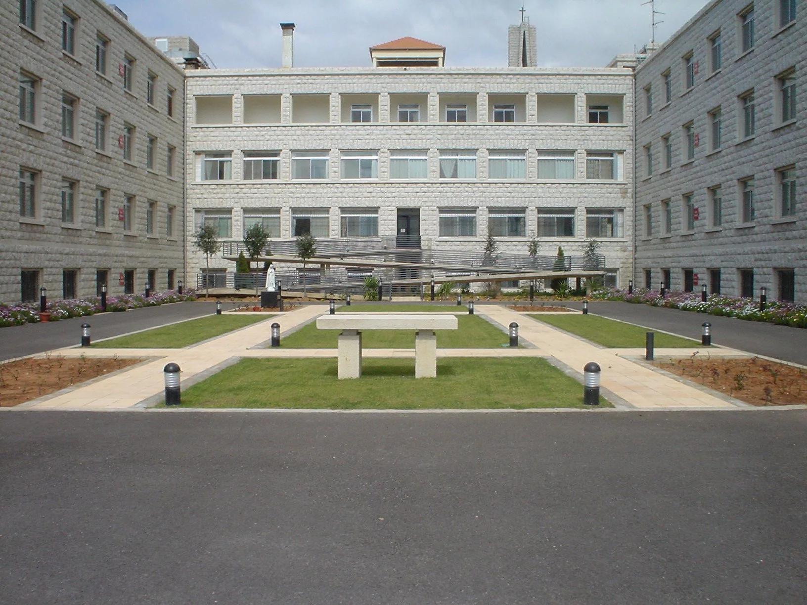 View of a modern apartment building with a landscaped courtyard featuring a concrete picnic table, pathways, flower beds, small trees, and decorative pathway lights.