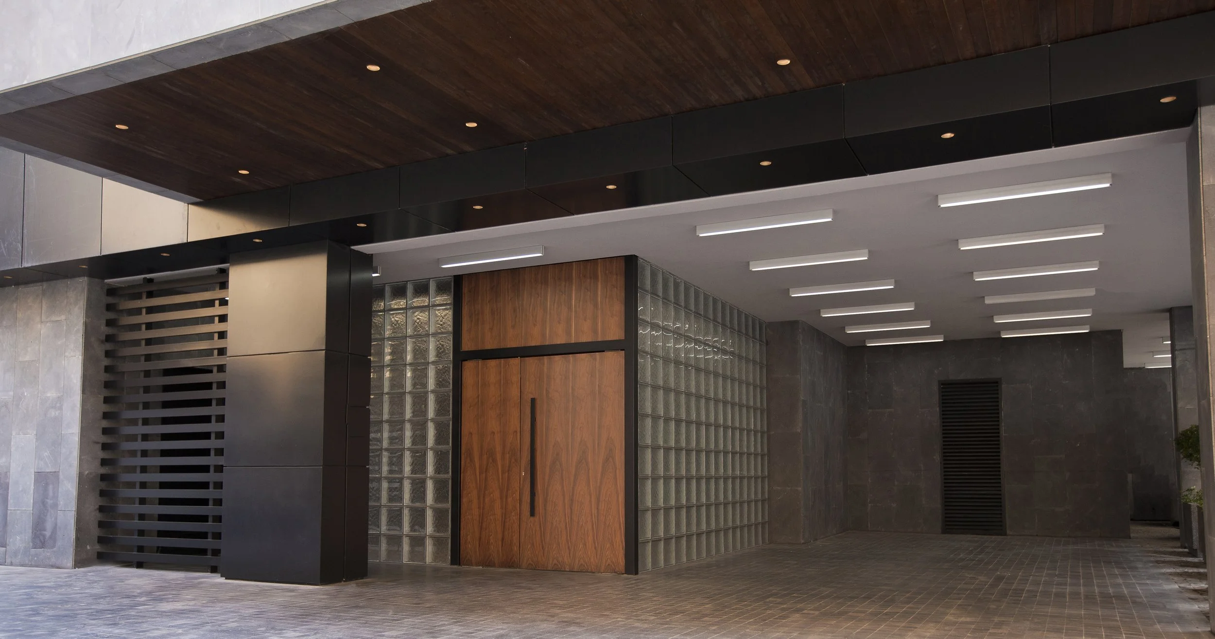 Modern building entrance with a wooden door, glass block wall, black wall paneling, and a ceiling with embedded lighting.