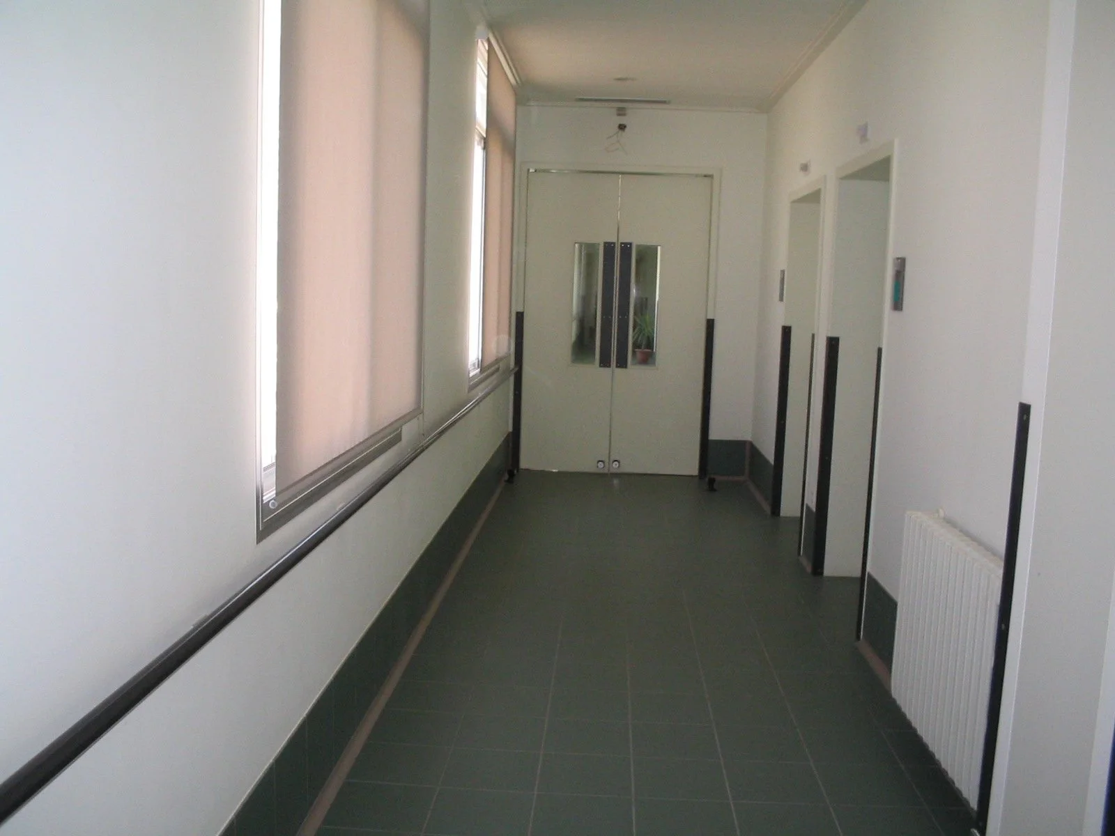 Empty hospital corridor with windows covered by blinds and two elevators with black strips on the right wall.