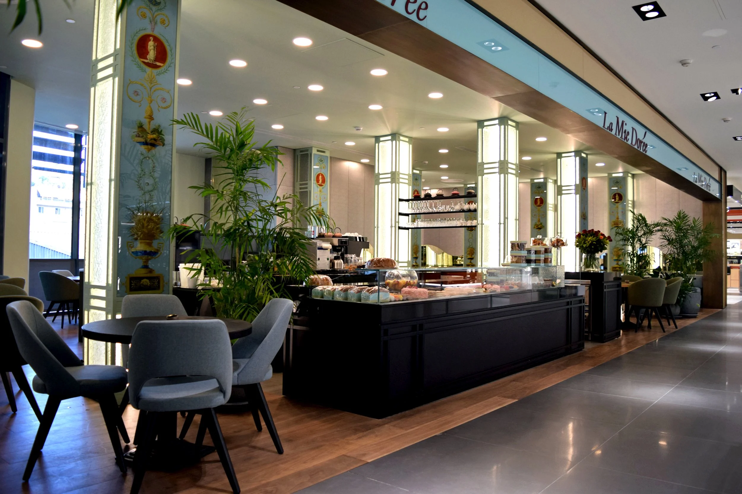 Interior of a cafe with gray chairs around small tables, green plants, display of pastries, and a counter with glassware and baked goods. Bright lighting and decorative columns inside a modern shopping mall or cafe.