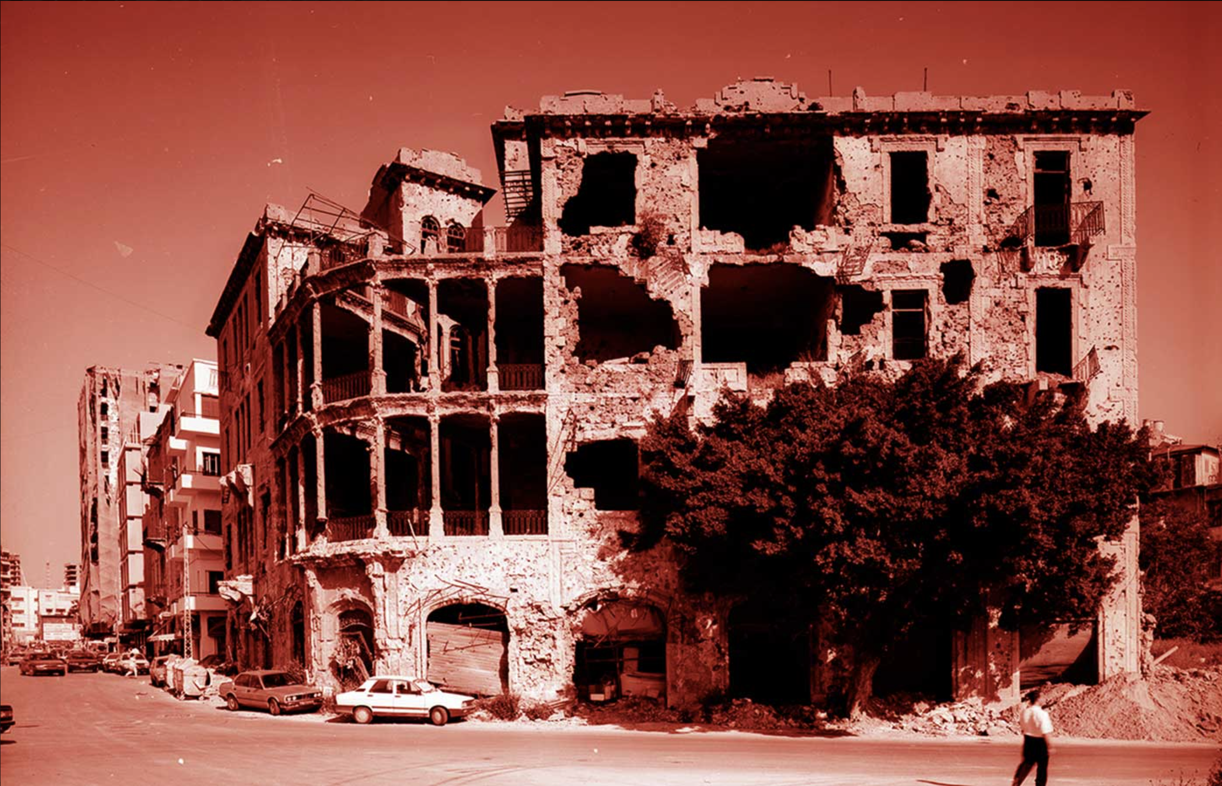 An old, partially ruined building with multiple floors and broken windows, situated in an urban area with cars parked nearby and a person walking in the foreground.