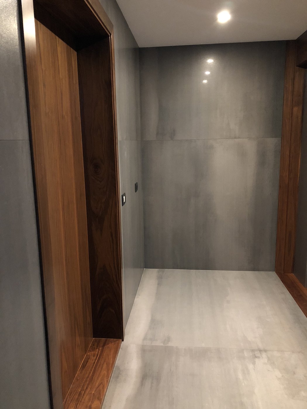 Empty elevator with grey walls, wooden door frame, and ceiling lights.