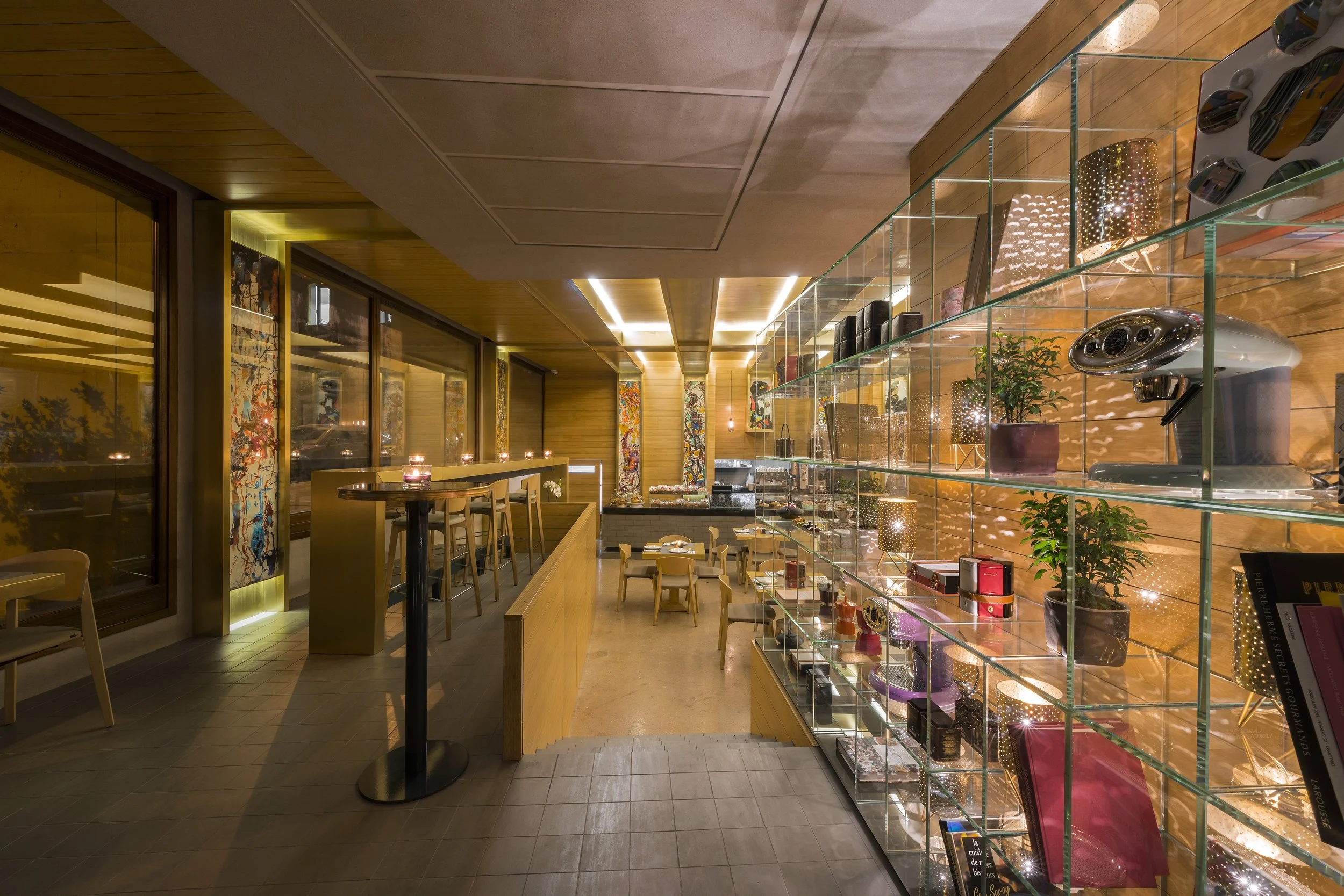 Interior of a modern restaurant or cafe with wooden walls and ceiling, glass shelving displaying books and decorative items, small tables with chairs, and warm lighting.