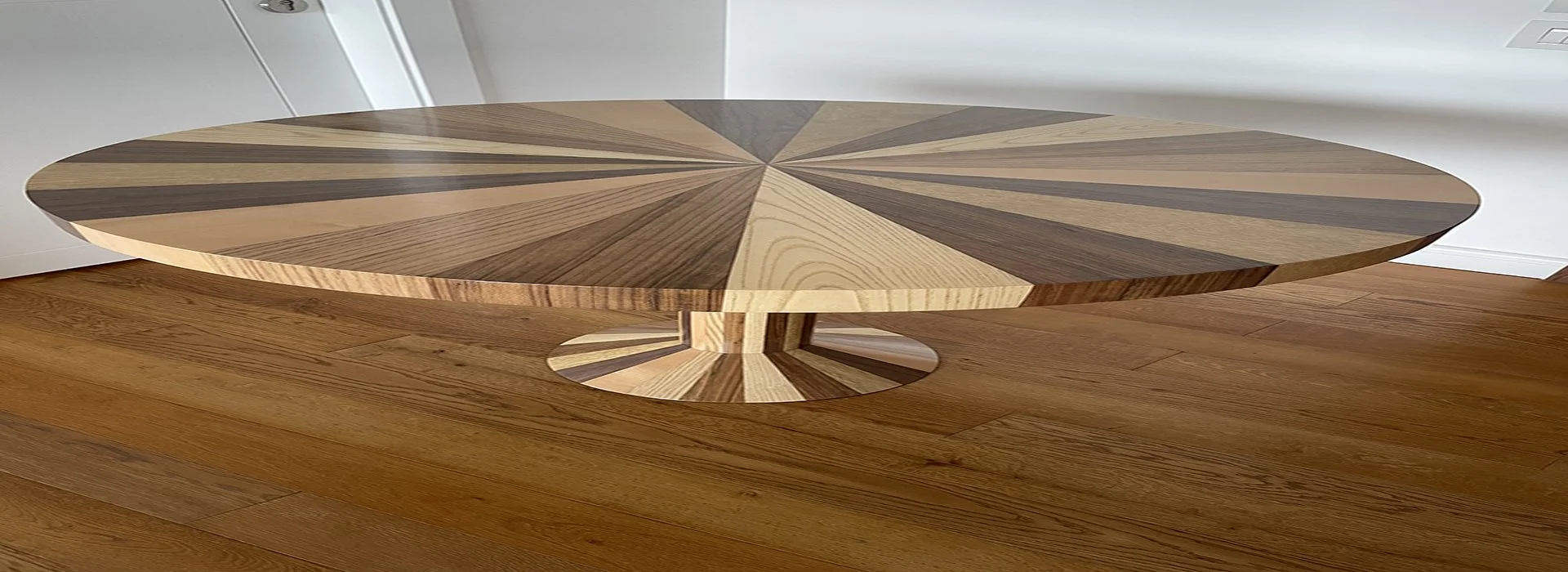 A round wooden dining table with a geometric pattern top, supported by a circular pedestal base with a matching pattern, placed on wooden flooring in a room with white walls.