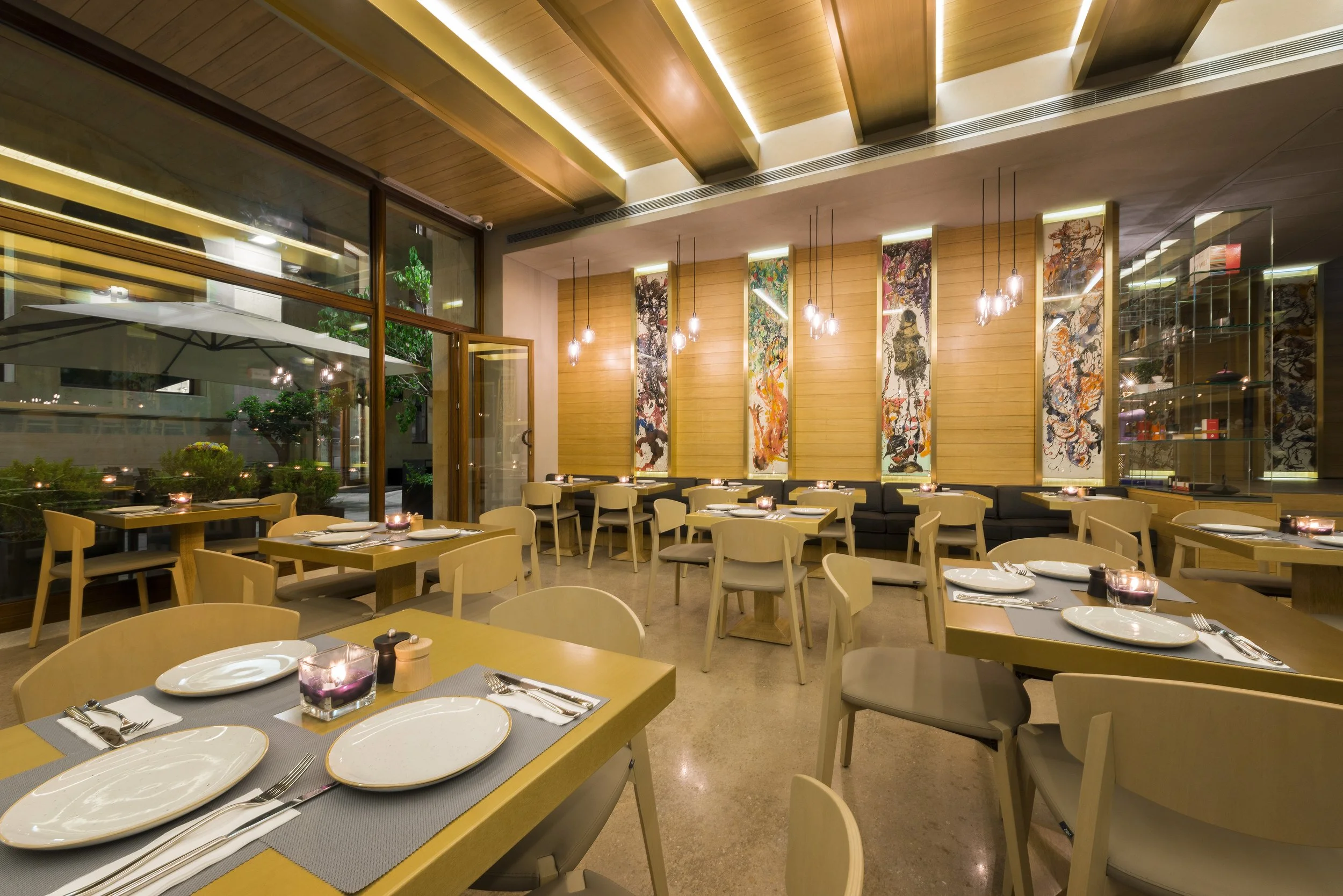 A modern restaurant interior with wooden ceiling beams, hanging pendant lights, large colorful abstract art panels on the wall, and a dining area with beige chairs and tables set with white plates, silverware, and candles in purple holders.