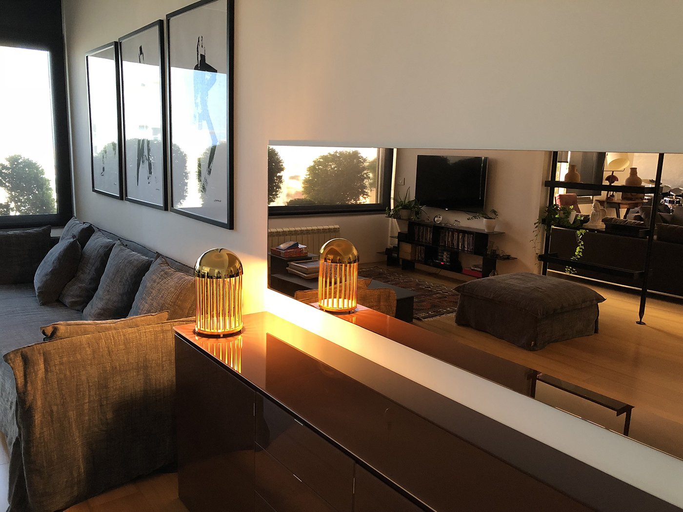 Living room with a sofa, artwork on the wall, and a mirror reflecting lamps and a TV, with sunlight outside the window.