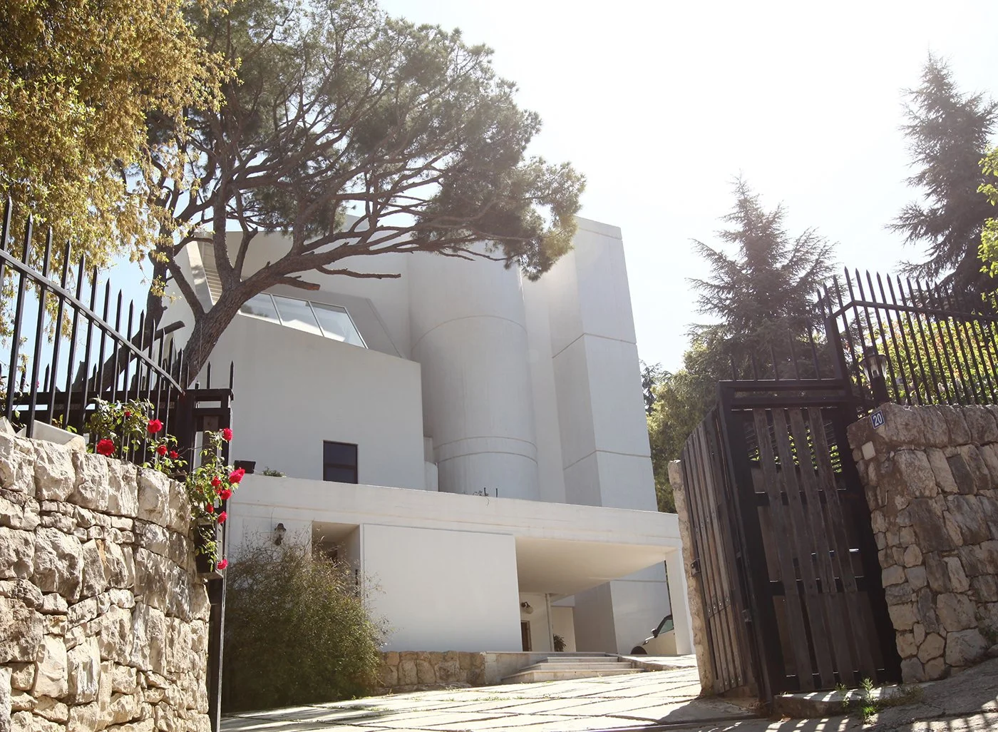 Modern white house with curved architecture, surrounded by stone and metal fencing, trees, and pink flowers, with sunlight brightening the scene.