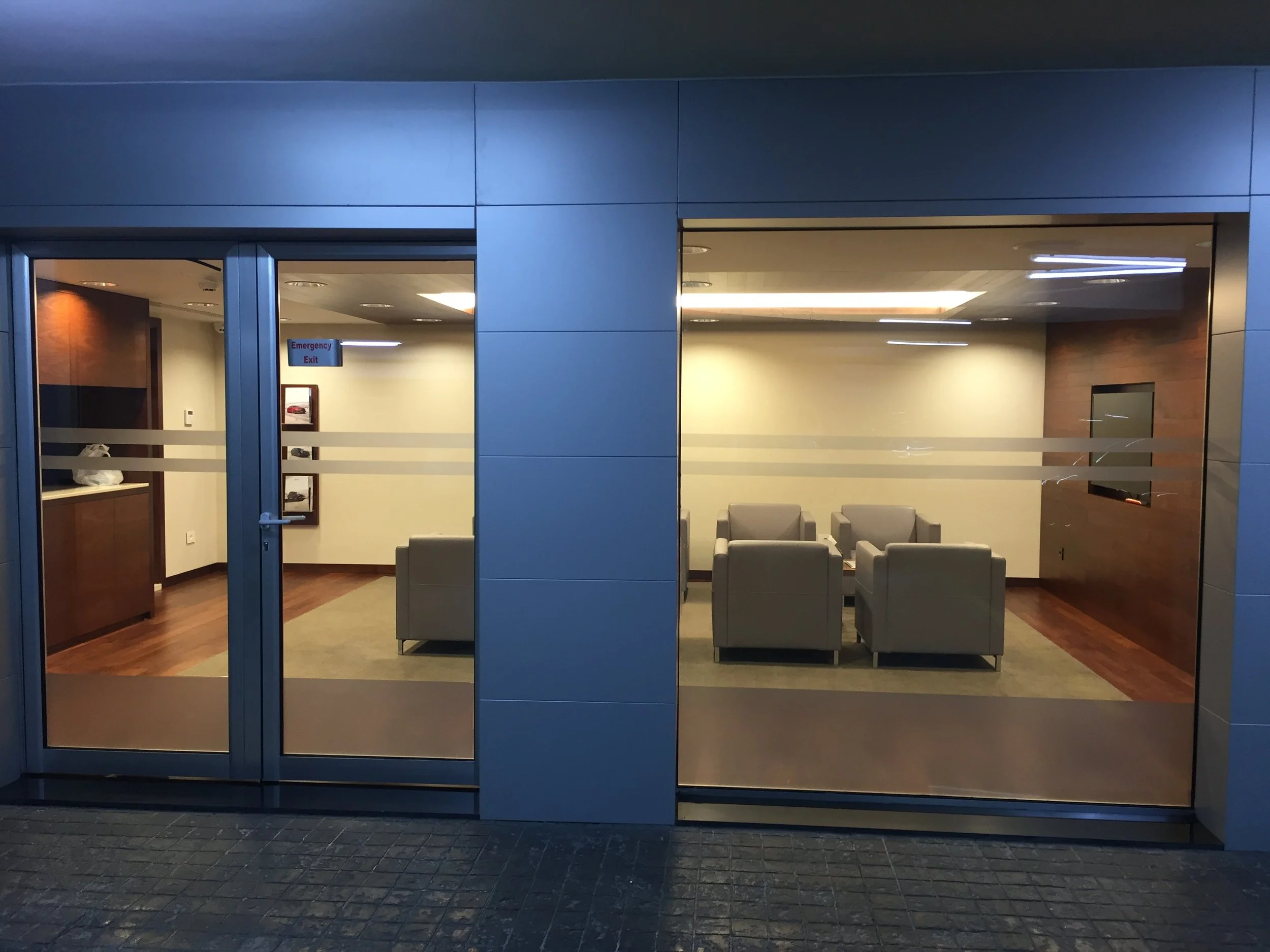 View of a modern waiting or lounge area with gray chairs arranged in a row, seen through large glass doors of a building at night. Inside, there is bright lighting, a sign indicating an emergency exit, and winter accessories on a table, with the inte