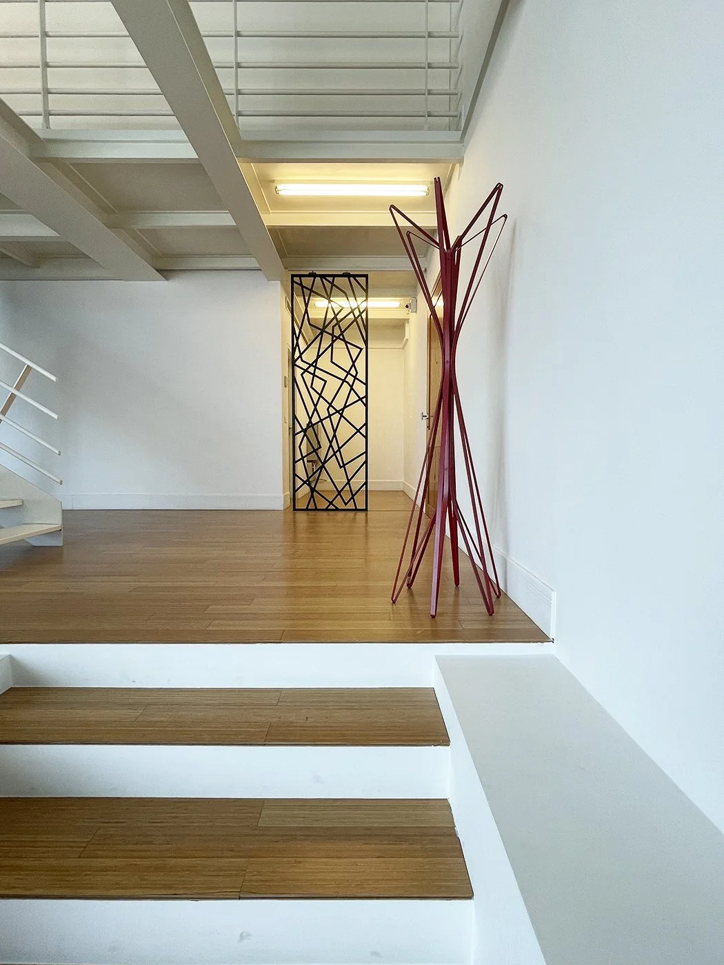 Interior view of a modern, minimalistic room with wooden steps, white walls, decorative red and black metal sculptures, and a patterned black metal screen in the background.