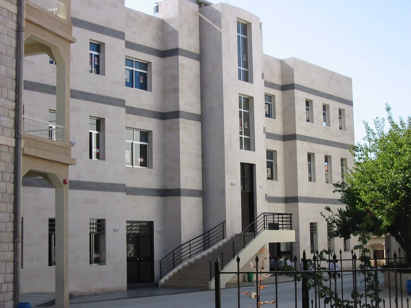 A modern multi-story building with beige and gray exterior walls and numerous windows, some with reflections of the sky and nearby objects. There is an outdoor staircase with black railings leading up to the entrance, and a small courtyard area with 