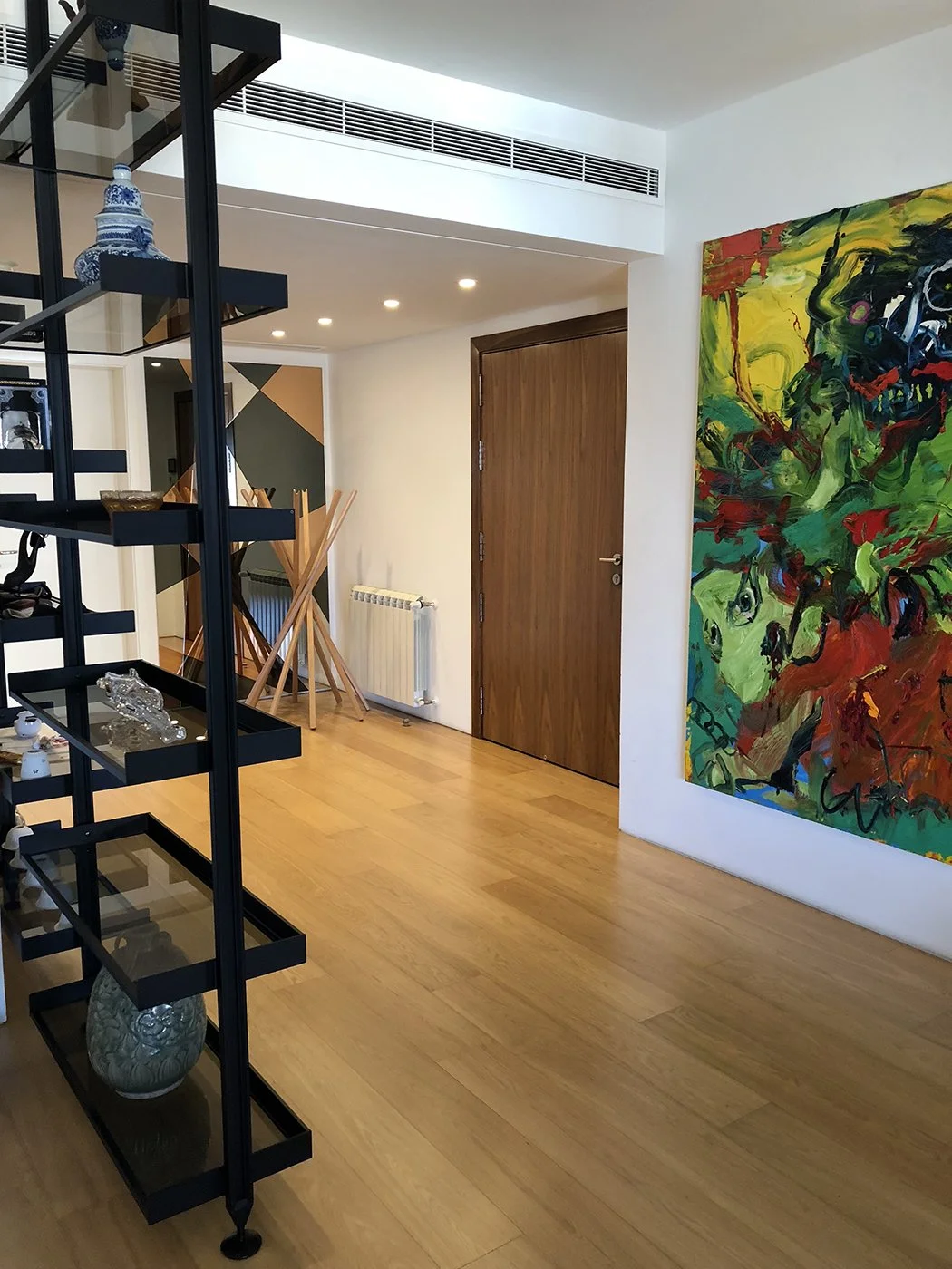 Modern interior living space with wooden flooring, a wooden door, a large colorful abstract painting on the wall, and black metal shelving unit holding decorative objects.